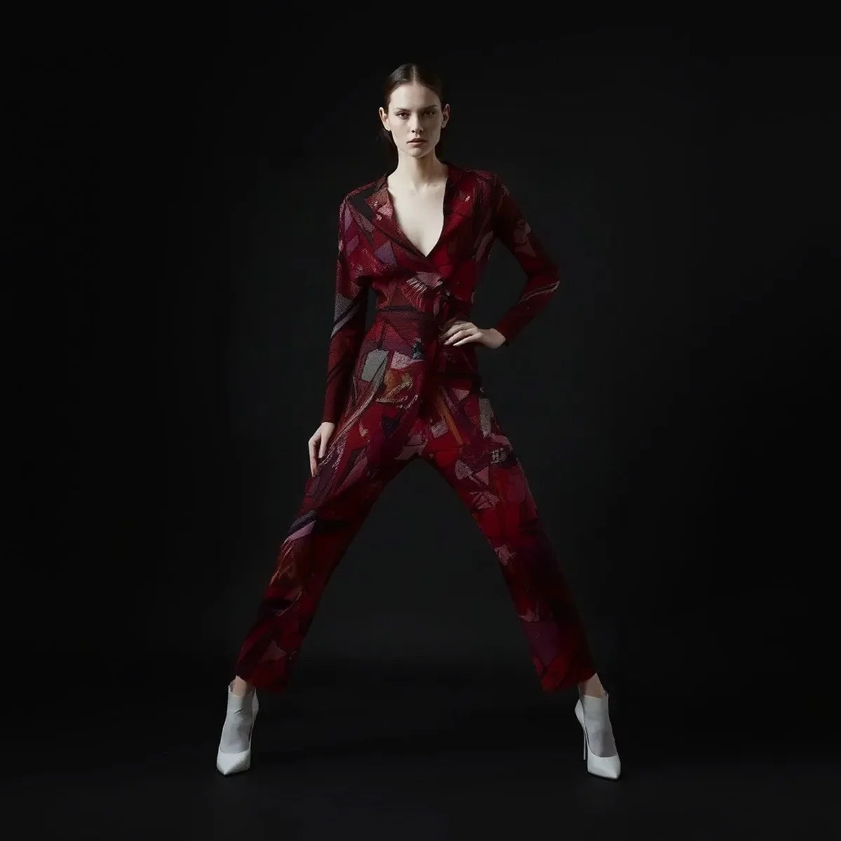 A woman in a red patterned pantsuit and white high-heeled shoes standing against a black background.