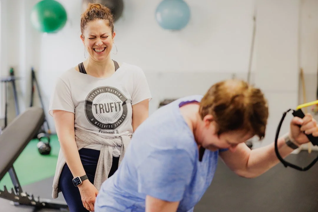 A woman laughing while working out with a trainer in a gym.