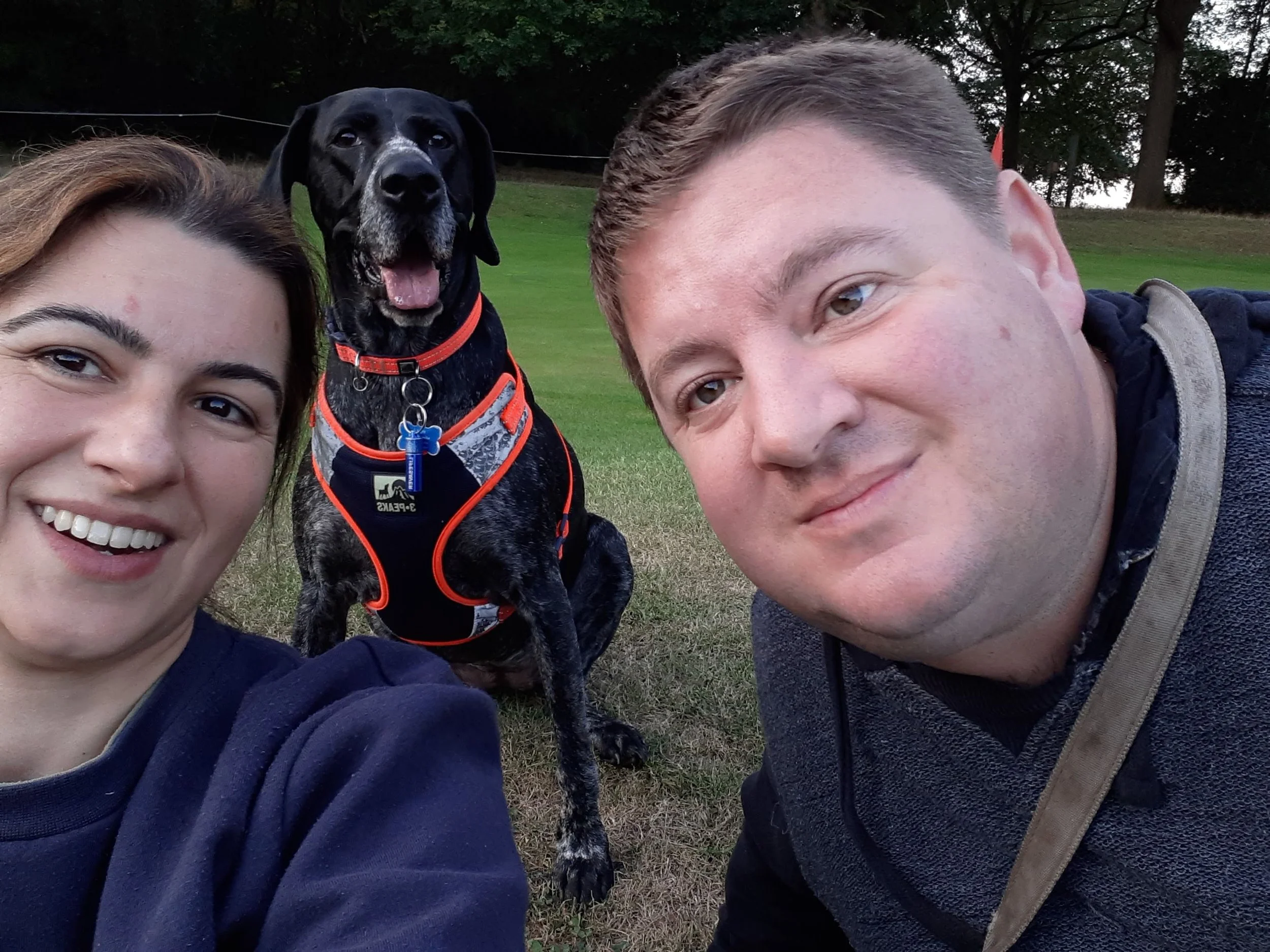 Two people taking a selfie with a black and white dog wearing an orange harness at a grassy park.