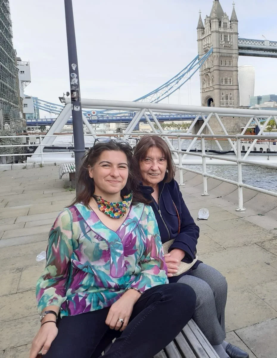 Two women sitting on a bench near the River Thames with the Tower Bridge in the background, in London, England.