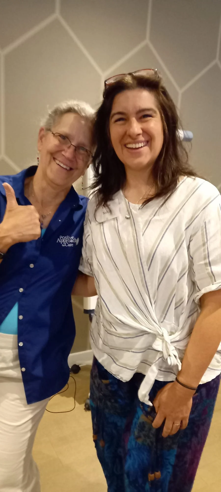 Two women smiling and posing for a selfie indoors. One woman is giving a thumbs-up, and the other woman is holding a phone.