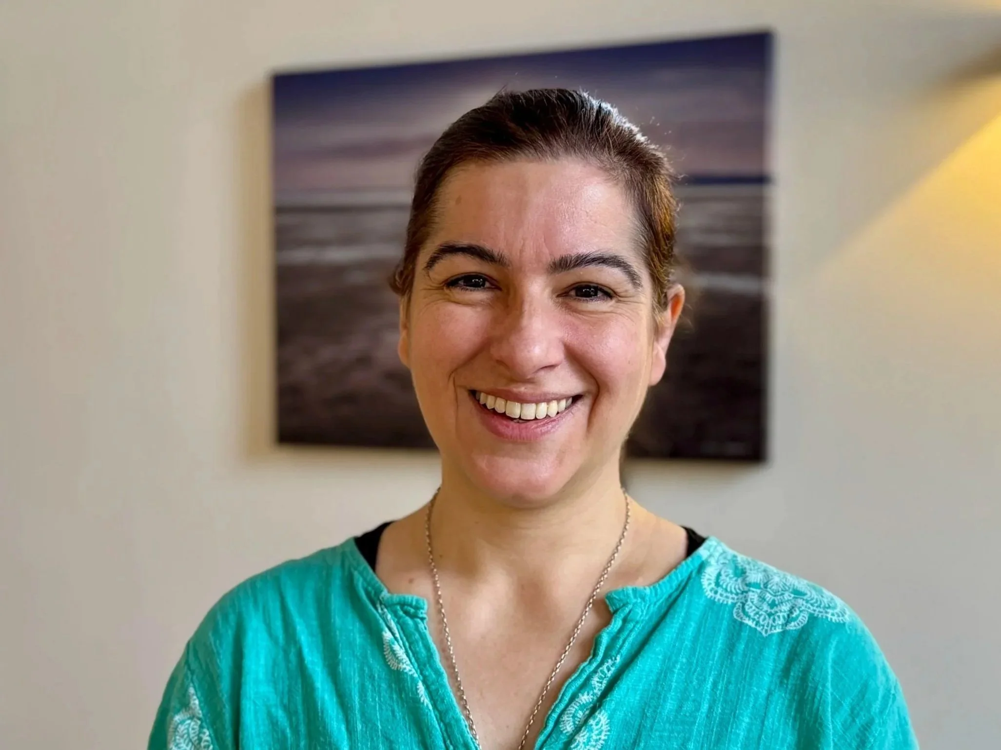 Smiling woman with brown hair tied back, wearing a turquoise shirt with embroidery, standing indoors with a framed picture of a seascape in the background.