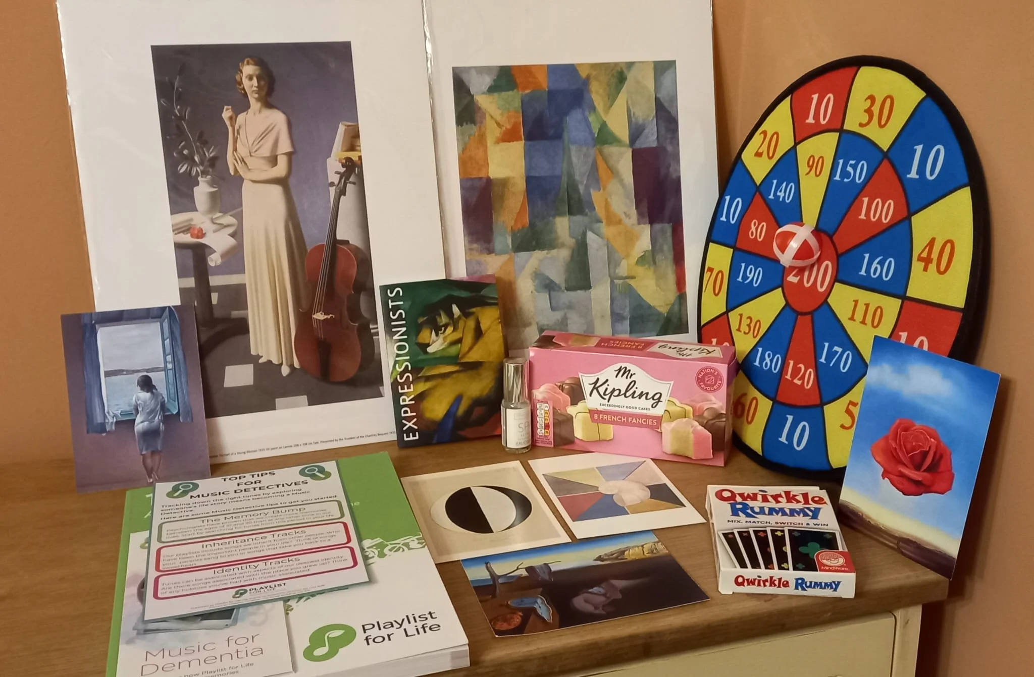 A collection of items on a table, including a woman with a cello, an abstract painting, a colorful wheel, a box of Mr. Kipling cakes, a pink card with a red rose, a quirkle rummy card game, a small bottle, and various printed materials about music and dementia.