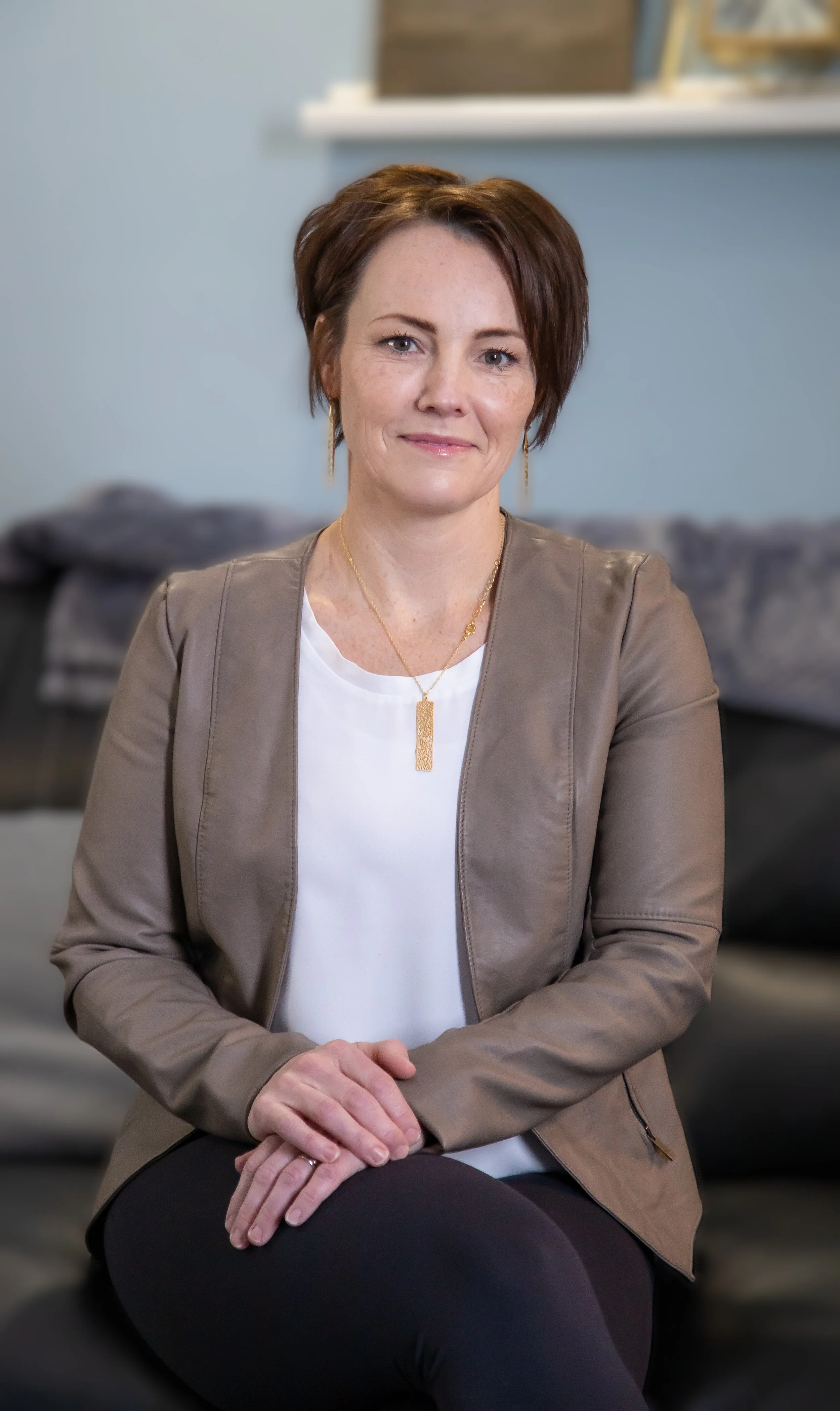 A woman with short brown hair sits with her hands clasped on her lap, wearing a tan jacket, white top, and gold jewelry, in a cozy living room with a blurred background.