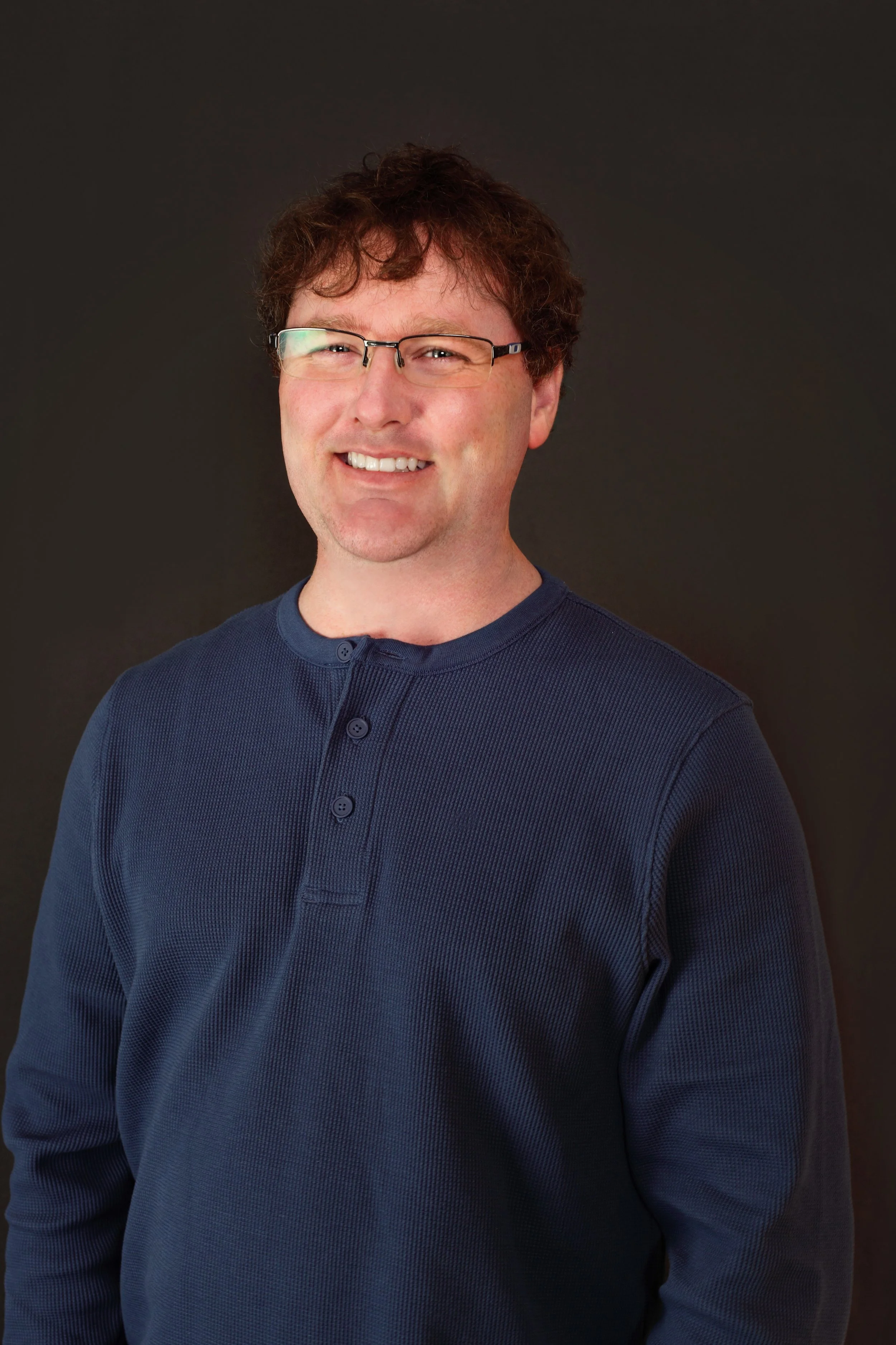 Portrait of a man with glasses, curly brown hair, wearing a dark green sweater, standing against a plain light-colored background.