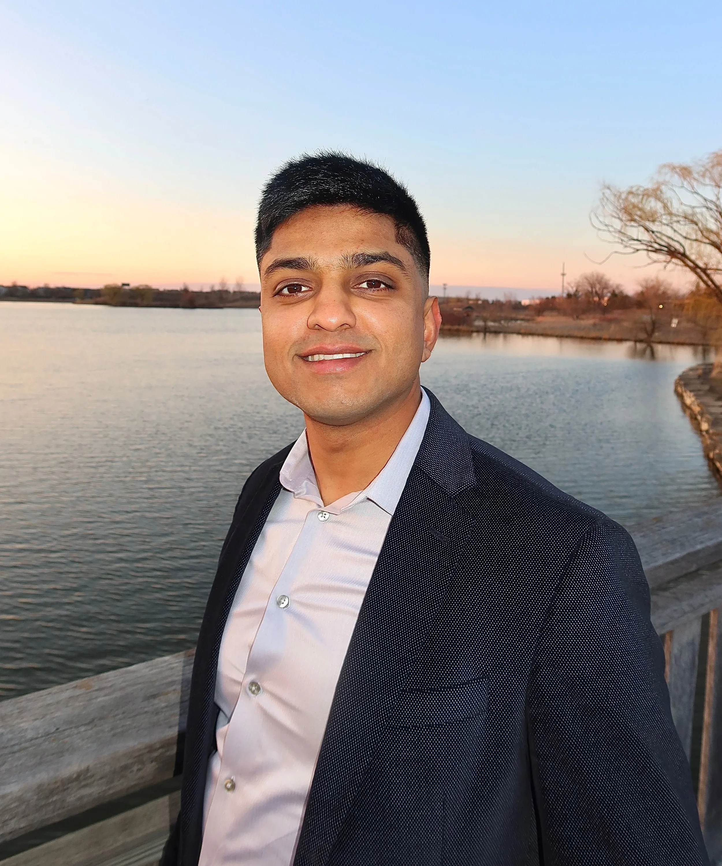 A young man with short black hair, wearing a dark blazer and white shirt, standing on a wooden dock by a body of water during sunset, with trees and a clear sky in the background.