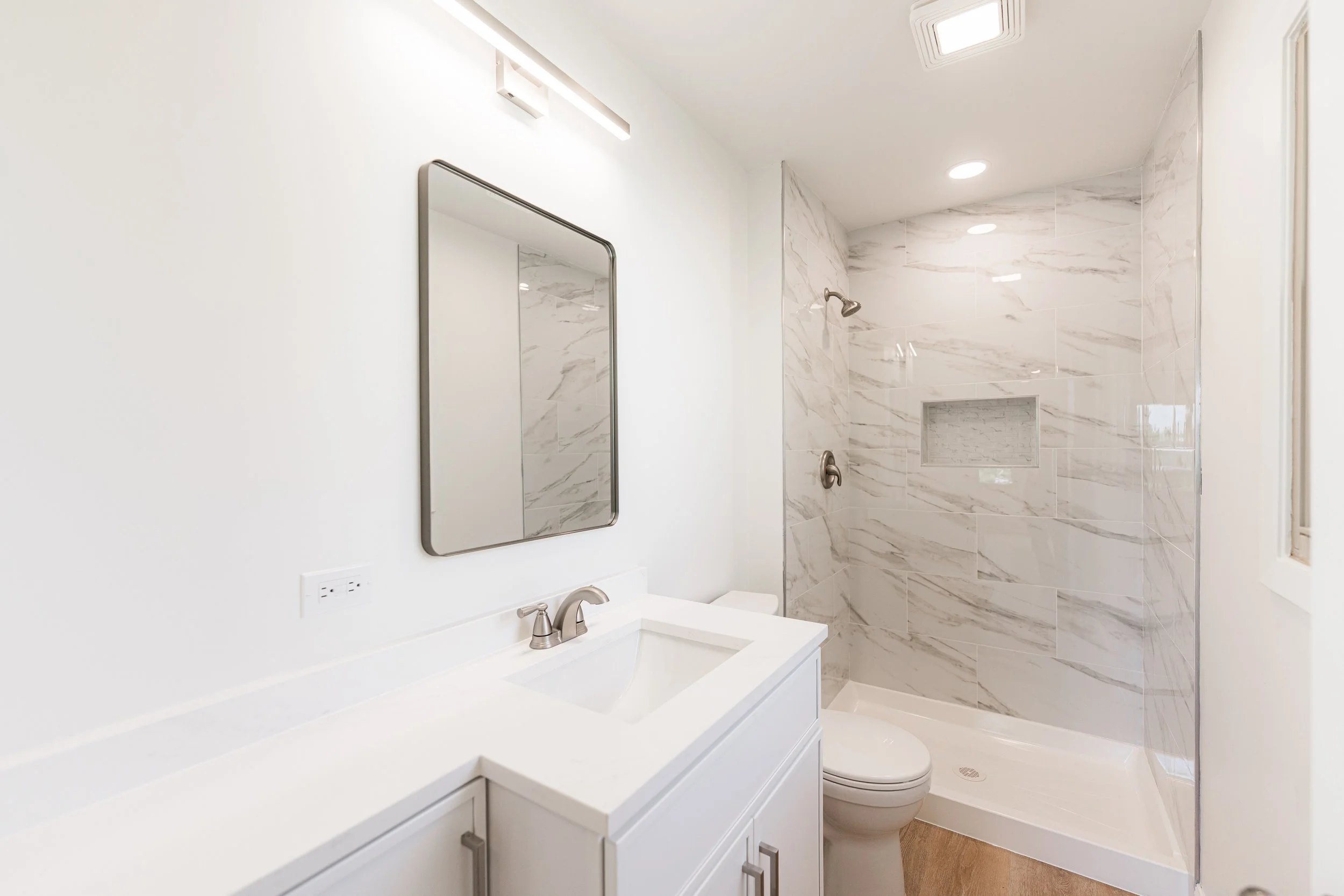 Modern bathroom with white cabinetry, a mirror, a white sink, a toilet, and a walk-in shower with marble walls and a built-in niche.