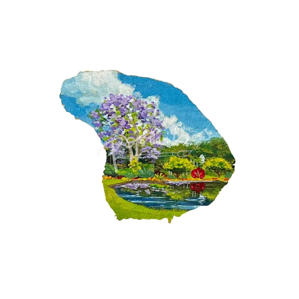 A hand-painted magnet in the shape of Lanai featuring a purple jacaranda tree in a lush garden.