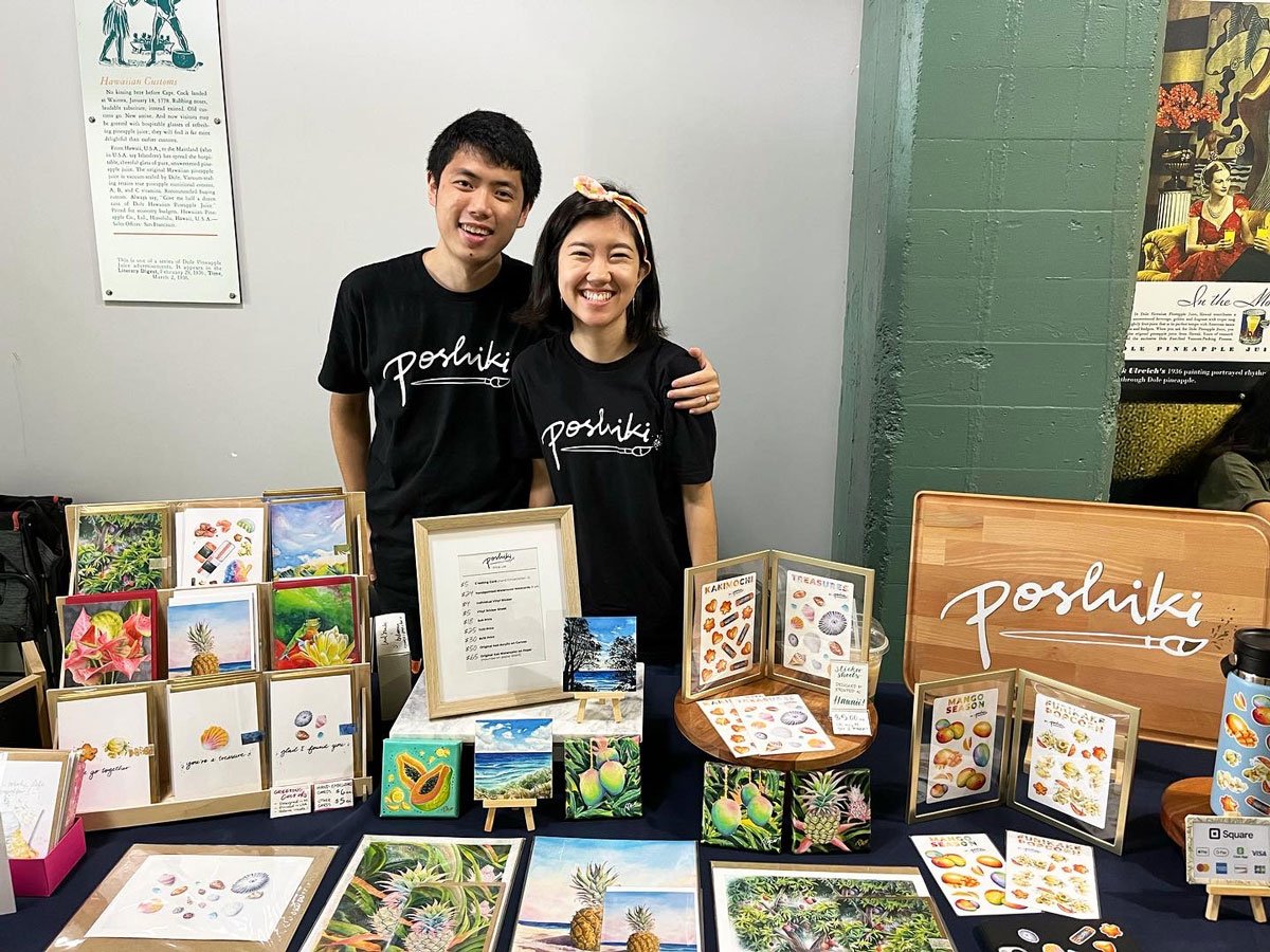 A husband and wife stand behind their art vendor table at a craft fair.