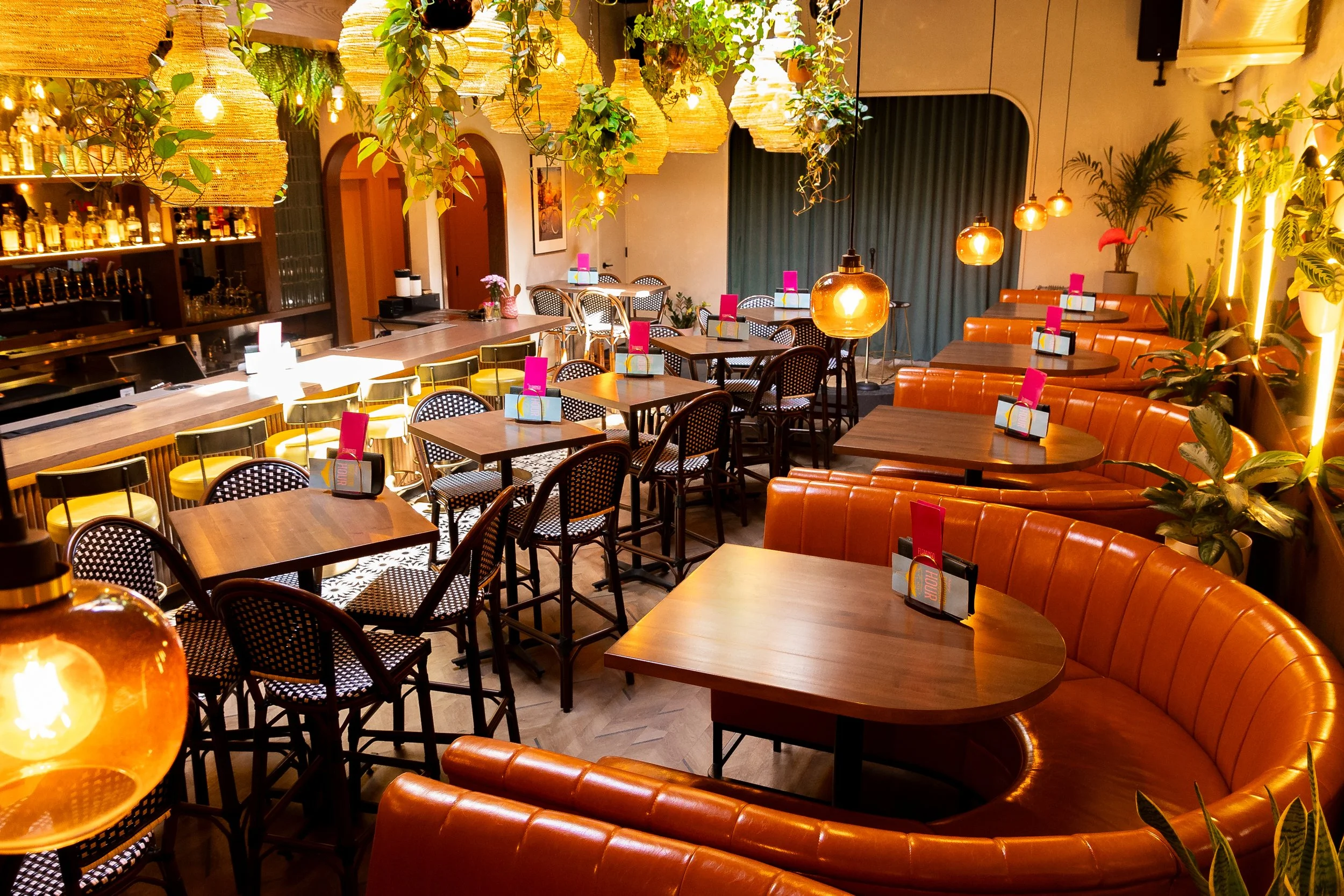 Interior of a stylish bar, the flamingo room with warm lighting, hanging woven lamps, potted plants, a bar counter on the left, and various tables and seating arrangements including orange curved booths and black-and-white patterned chairs.