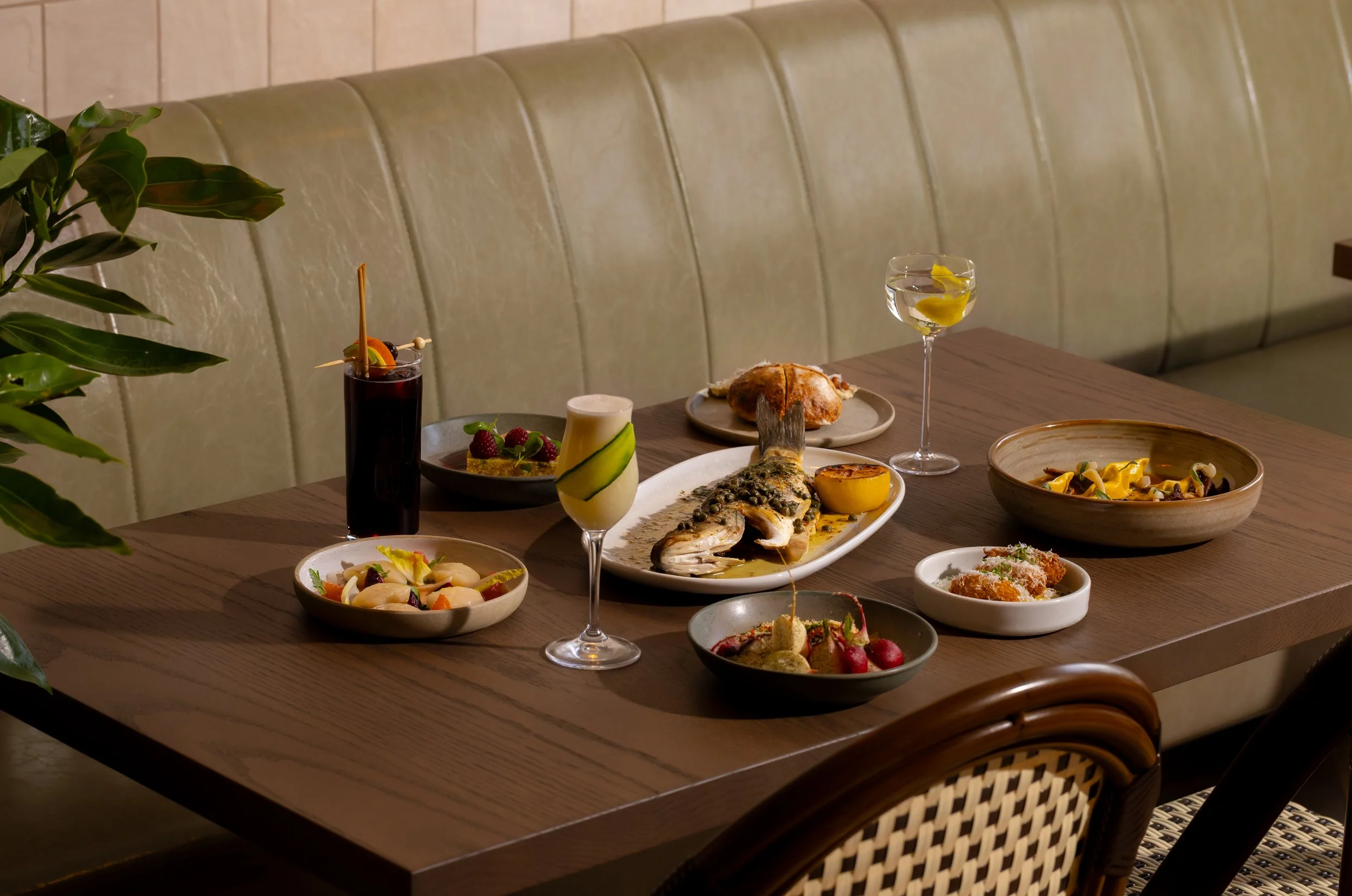 A dining table set with various dishes including whole fish with lemon, pasta, salads, and desserts, along with cocktails and drinks, in an elegant restaurant setting.