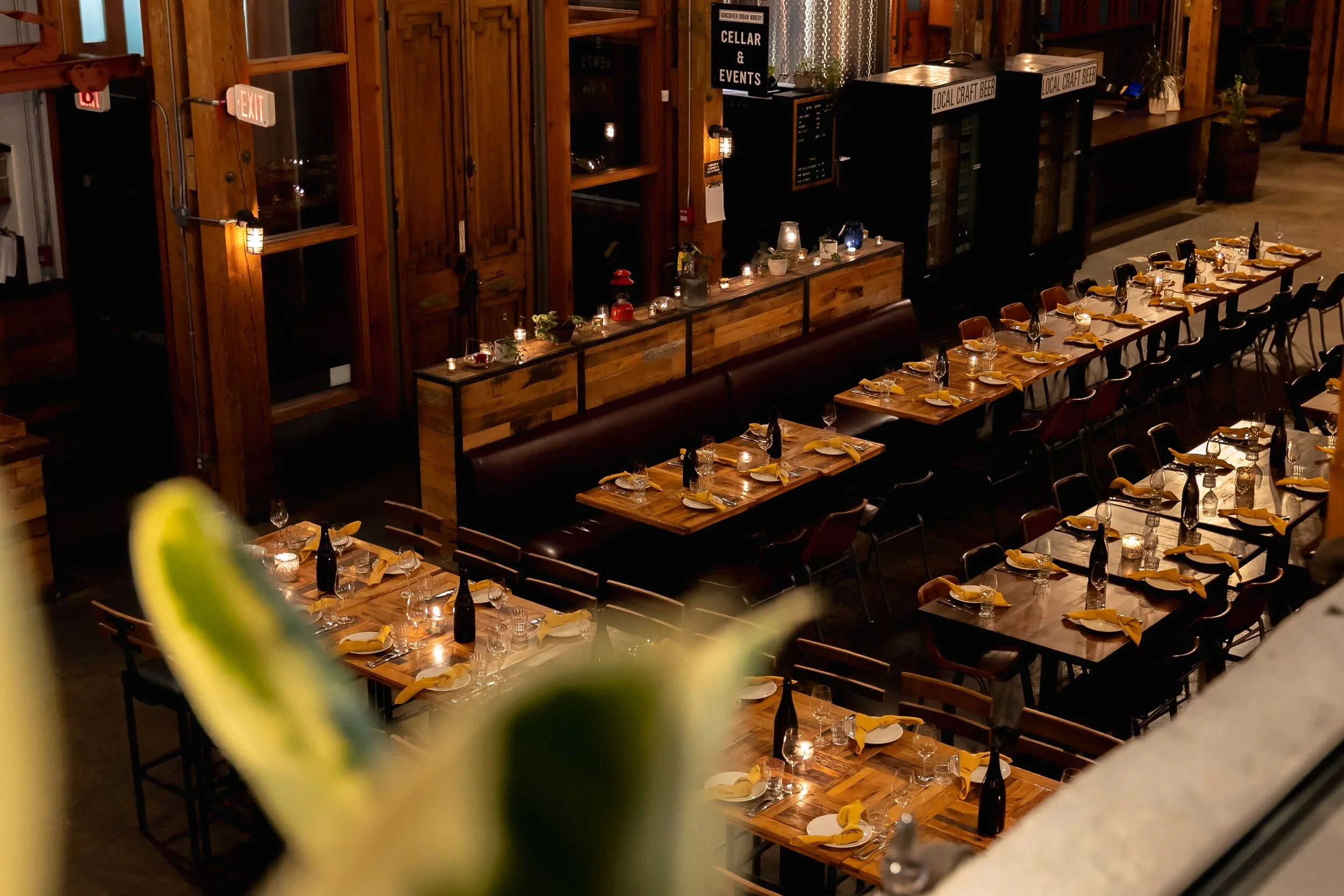 Vancouver urban winery Interior of a restaurant with wooden tables set for dining, wine bottles, napkins, and glasses, and warm ambient lighting.