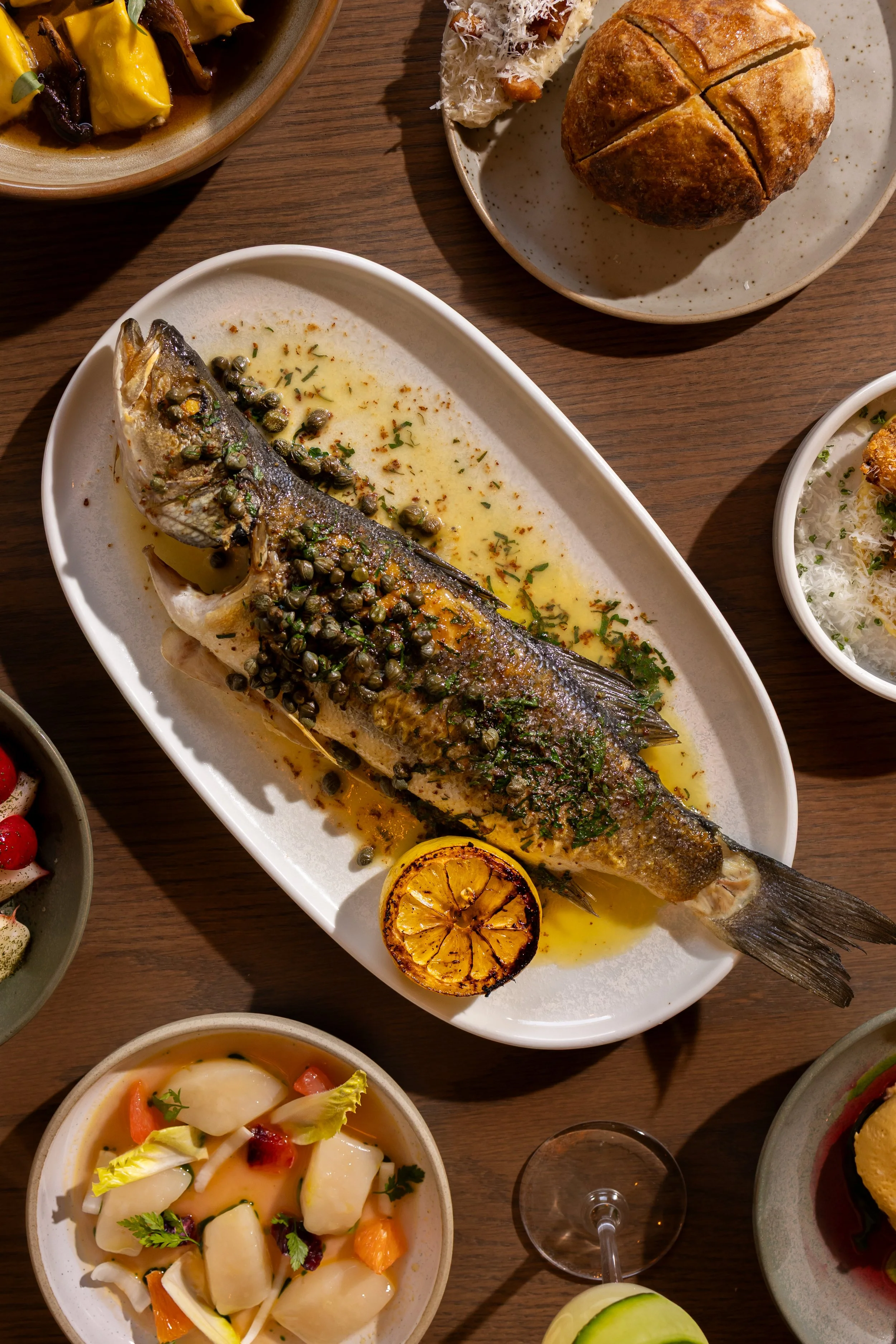 A whole cooked fish topped with herbs and capers on a white platter, garnished with a grilled lemon slice, surrounded by various side dishes including bread, salad, and other appetizers on a wooden table.