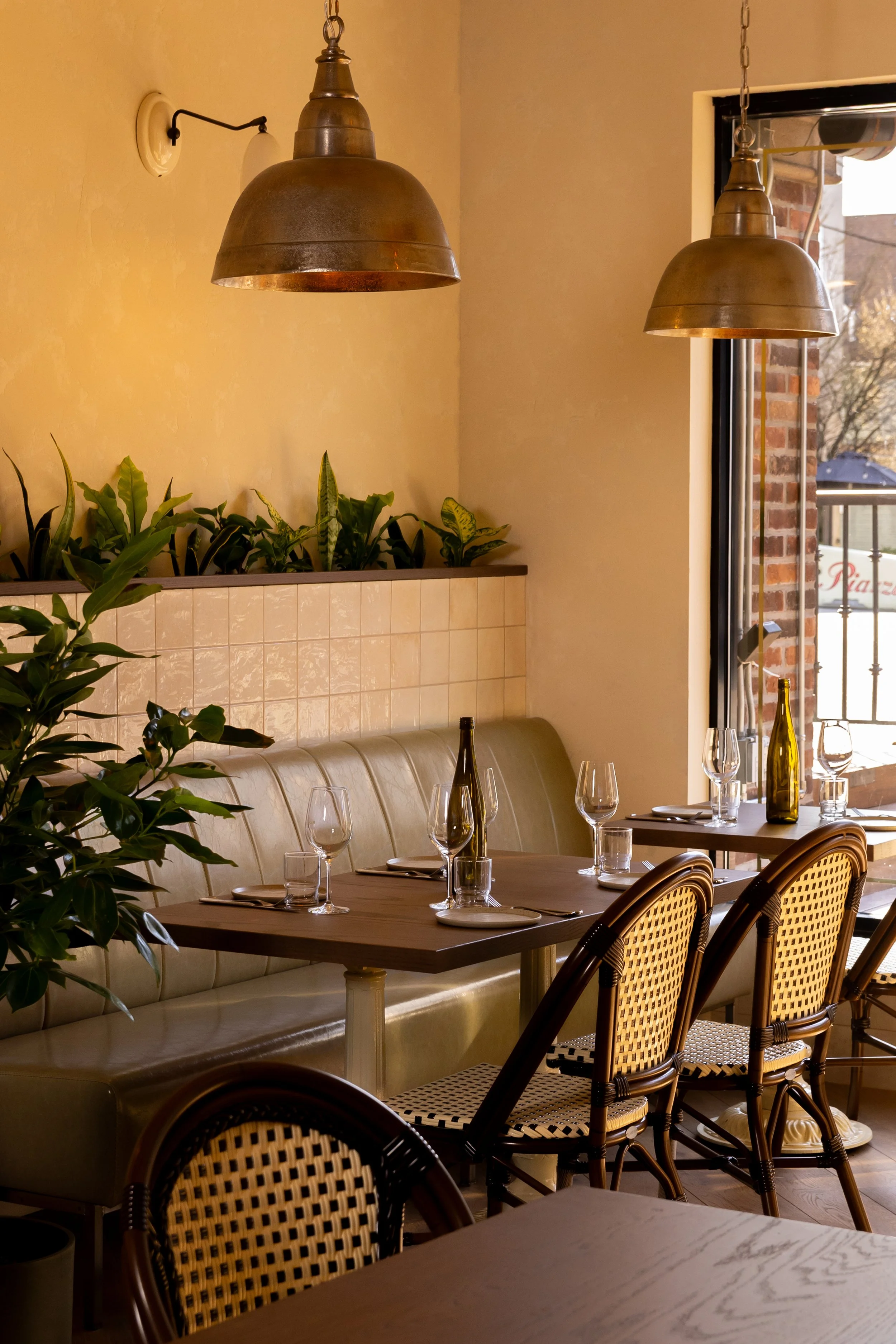 Interior of a cozy restaurant with a dining table set with wine glasses, a bottle of wine, and decorative bottles. There are rattan chairs, a beige leather bench, greenery, warm lighting, and a window showing an outdoor scene.