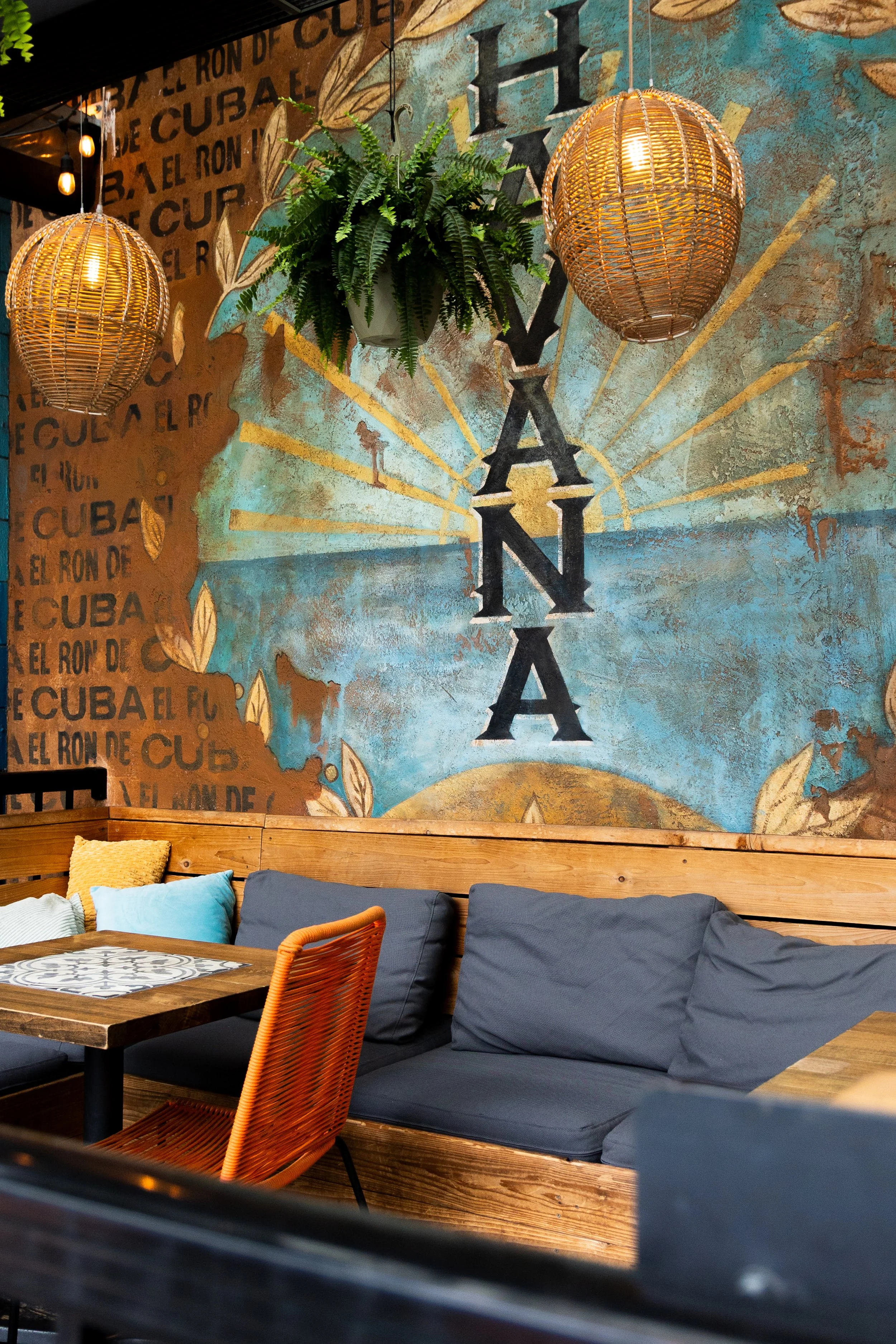 Cozy restaurant exterior with a colorful wall mural, including the word 'HAVANA' with a sunrise design, hanging wicker lamps, a wooden bench with gray cushions, pillows, a wooden table, and a potted fern plant.