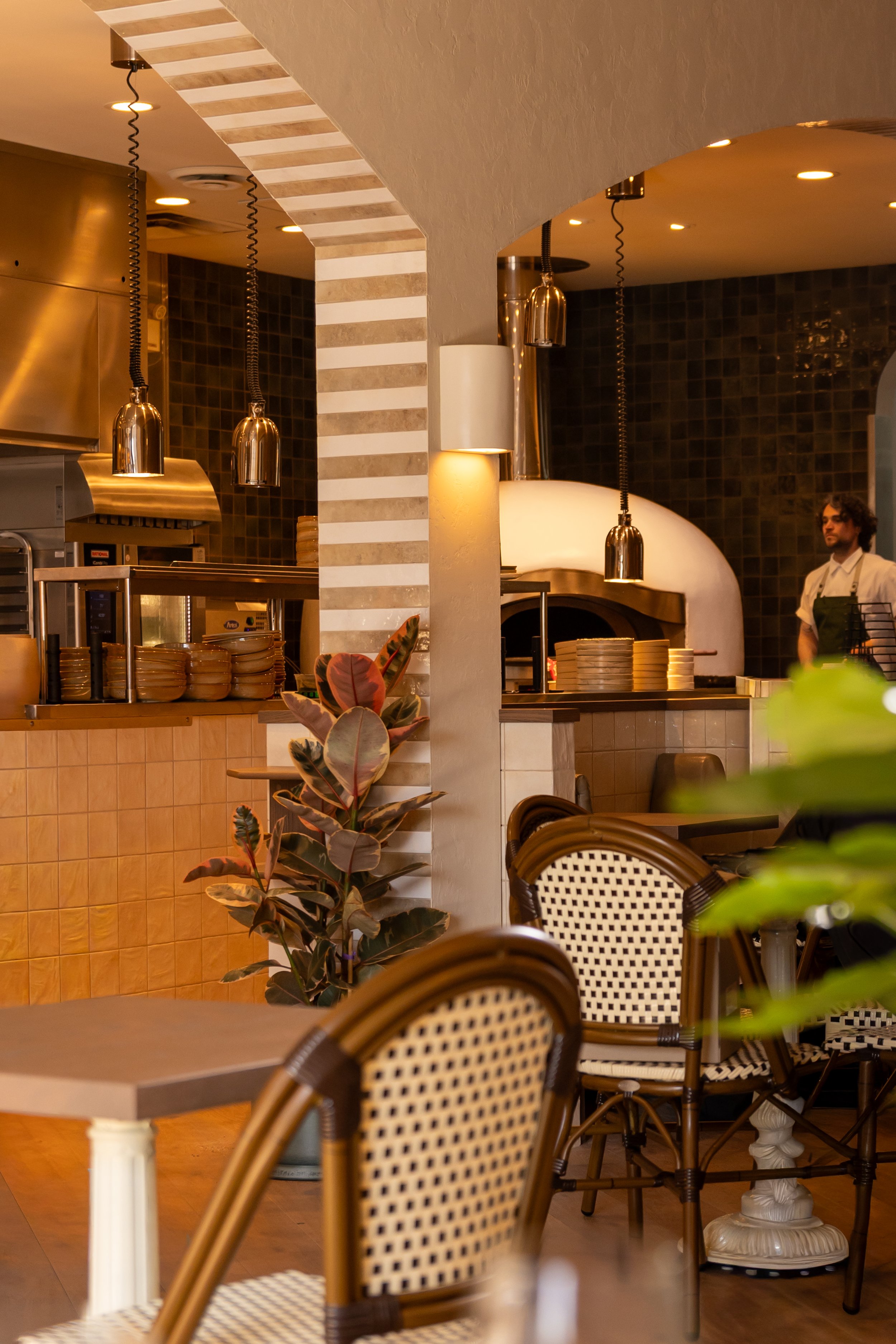 Inside a cozy restaurant with a partial view of the kitchen, wicker chairs with cream-colored cushions, indoor plants, and warm lighting.