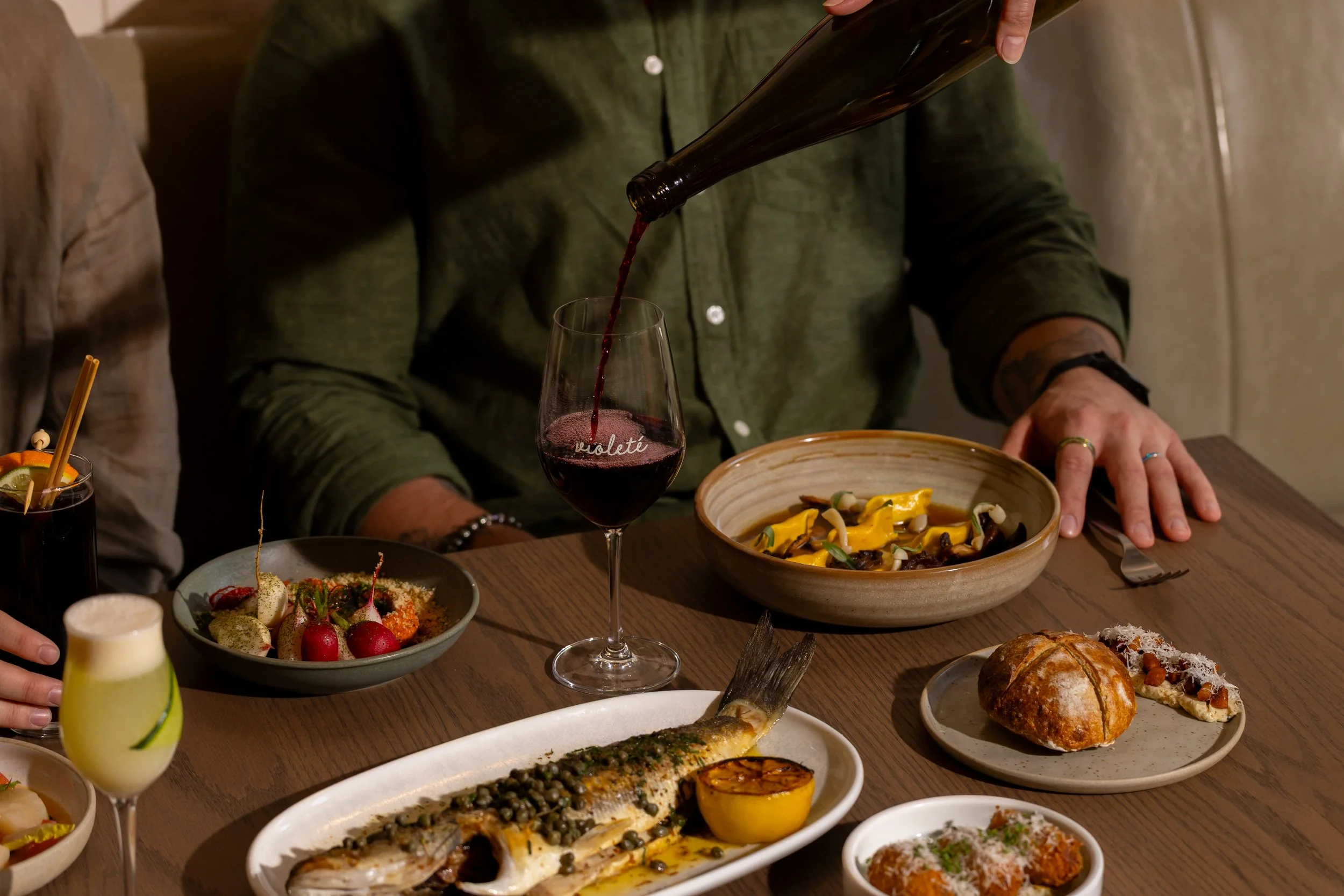 People dining at a restaurant table with various dishes, wine, and cocktails.