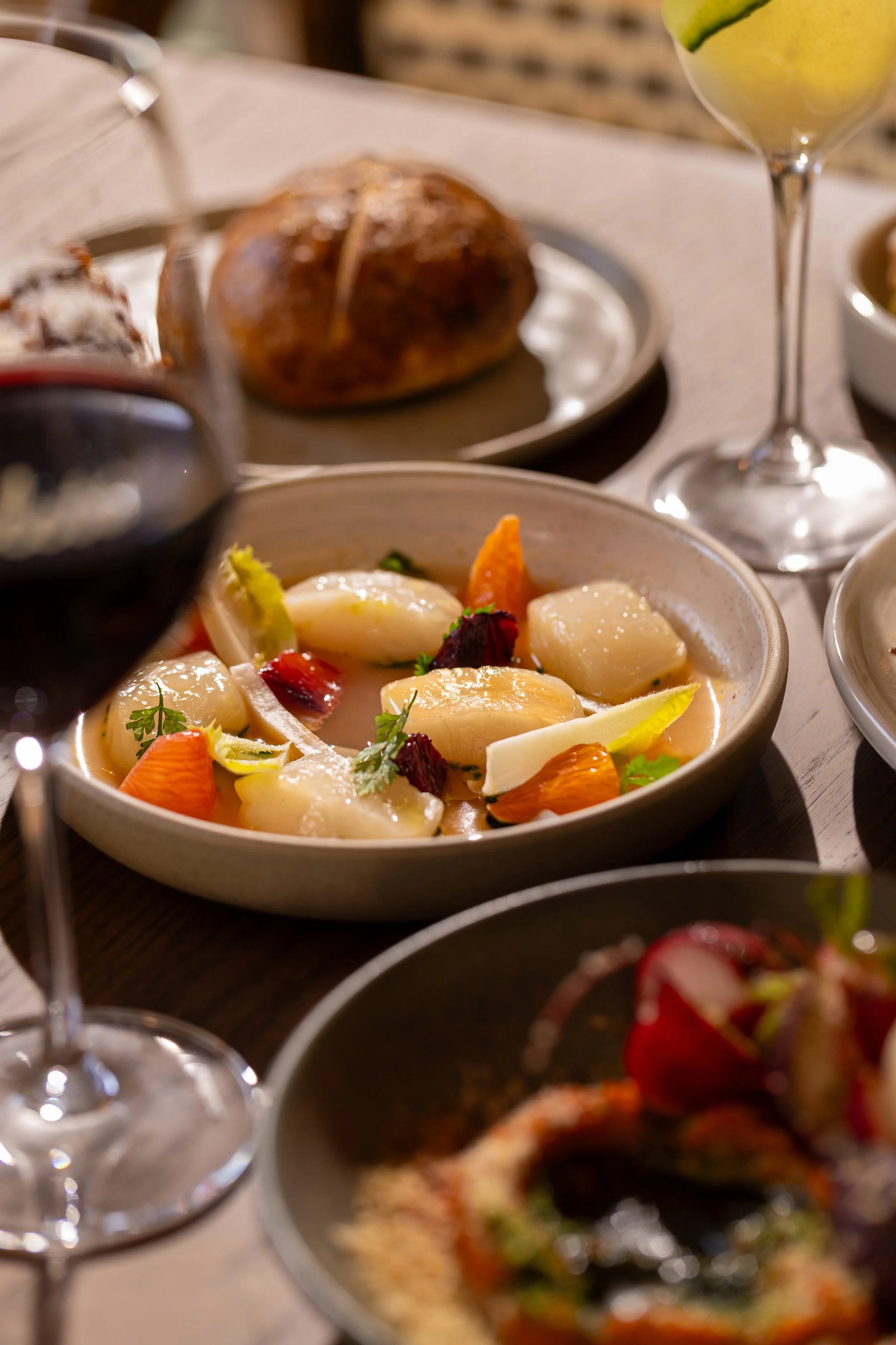 A bowl of scallops on a table dinner setting, with wine glasses and a plate of bread in the background.