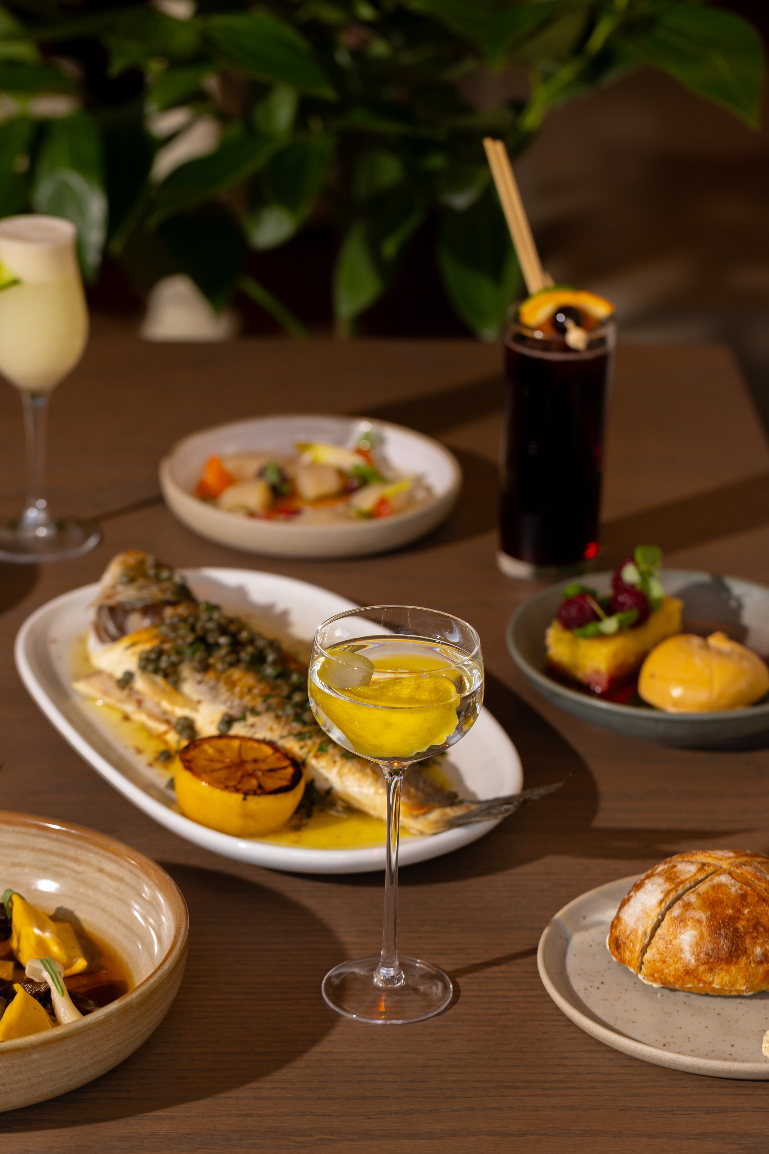 A variety of dishes and drinks on a wooden table, including a grilled fish with lemon, a glass of white wine, a dark cocktail with garnish, a small dessert with berries, and a bread roll.