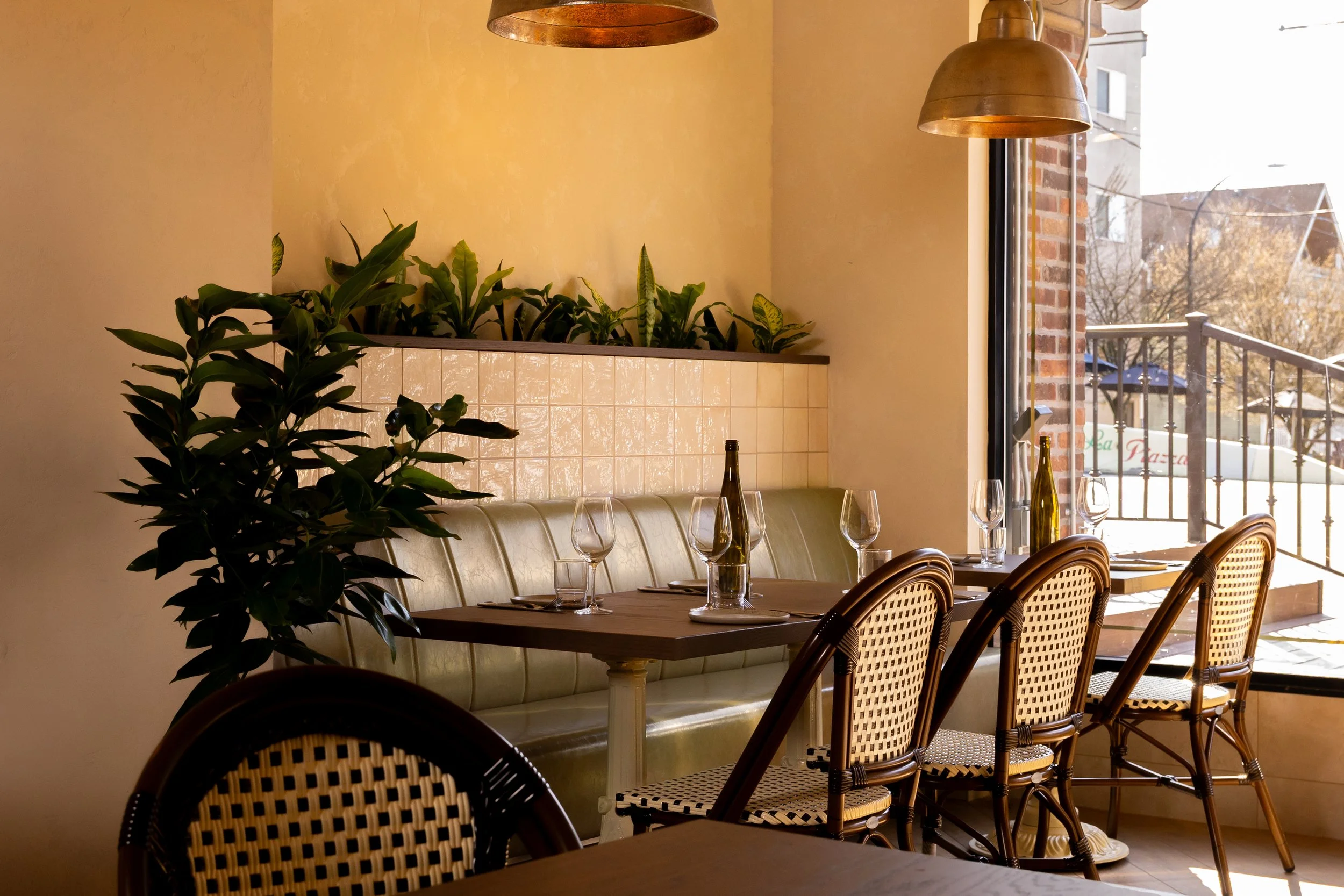 Empty restaurant table set with glasses and bottles near a large window, with a beige cushioned bench and wicker chairs, sunlight streaming in, and indoor plants.