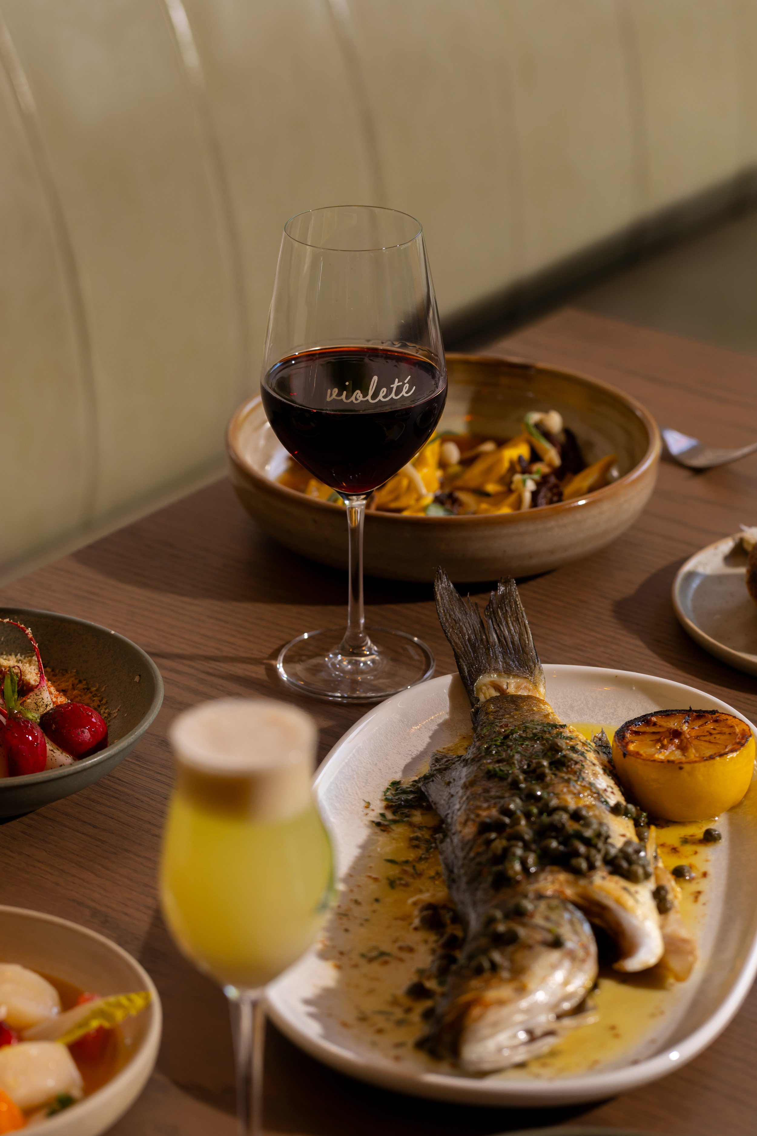 A table with a grilled fish served on a white platter with lemon, a glass of red wine labeled "violeté", a glass of yellow-green beverage, a bowl of pasta, and small dishes with cherry tomatoes and other appetizers.