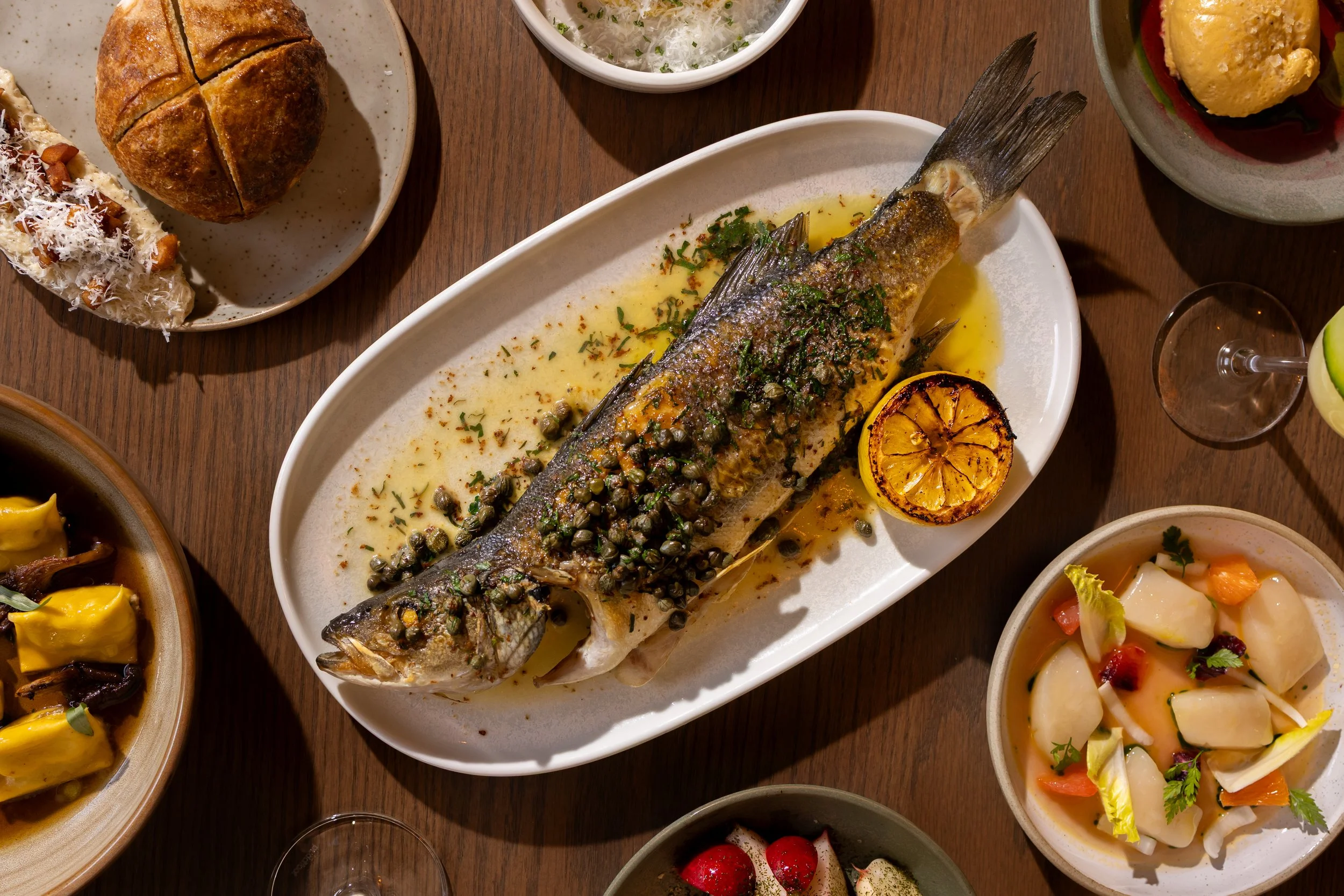 Whole grilled fish topped with herbs and capers, garnished with grilled lemon half, surrounded by various side dishes on a wooden table.