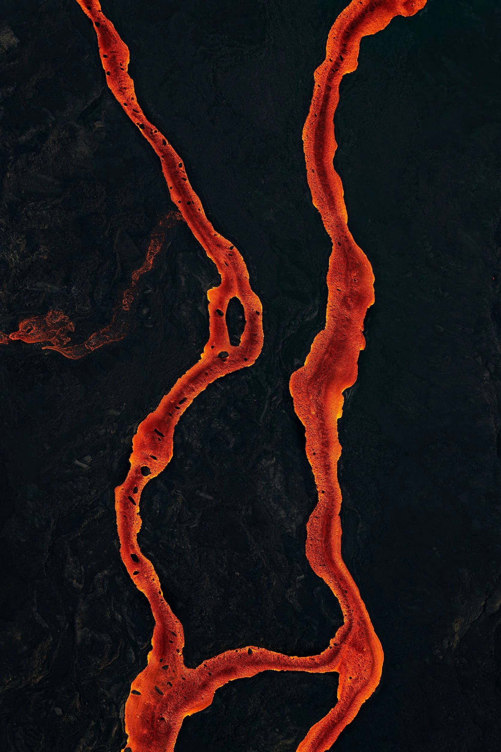 Aerial view of a dark landscape with bright orange lava streams flowing across, creating winding paths.