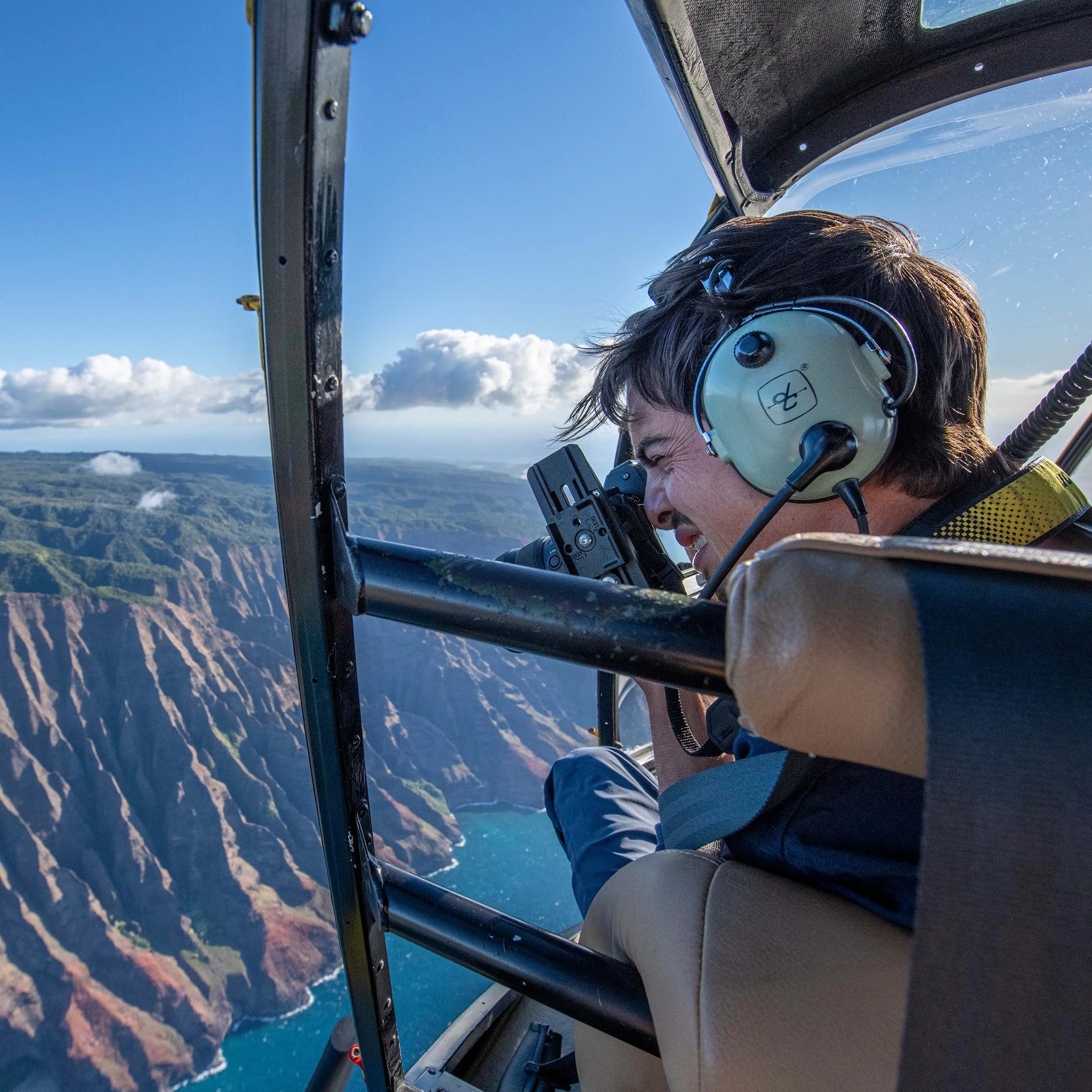 Person inside an airplane cockpit, wearing headphones and looking through a camera, flying over a landscape of cliffs and water under a blue sky with clouds.
