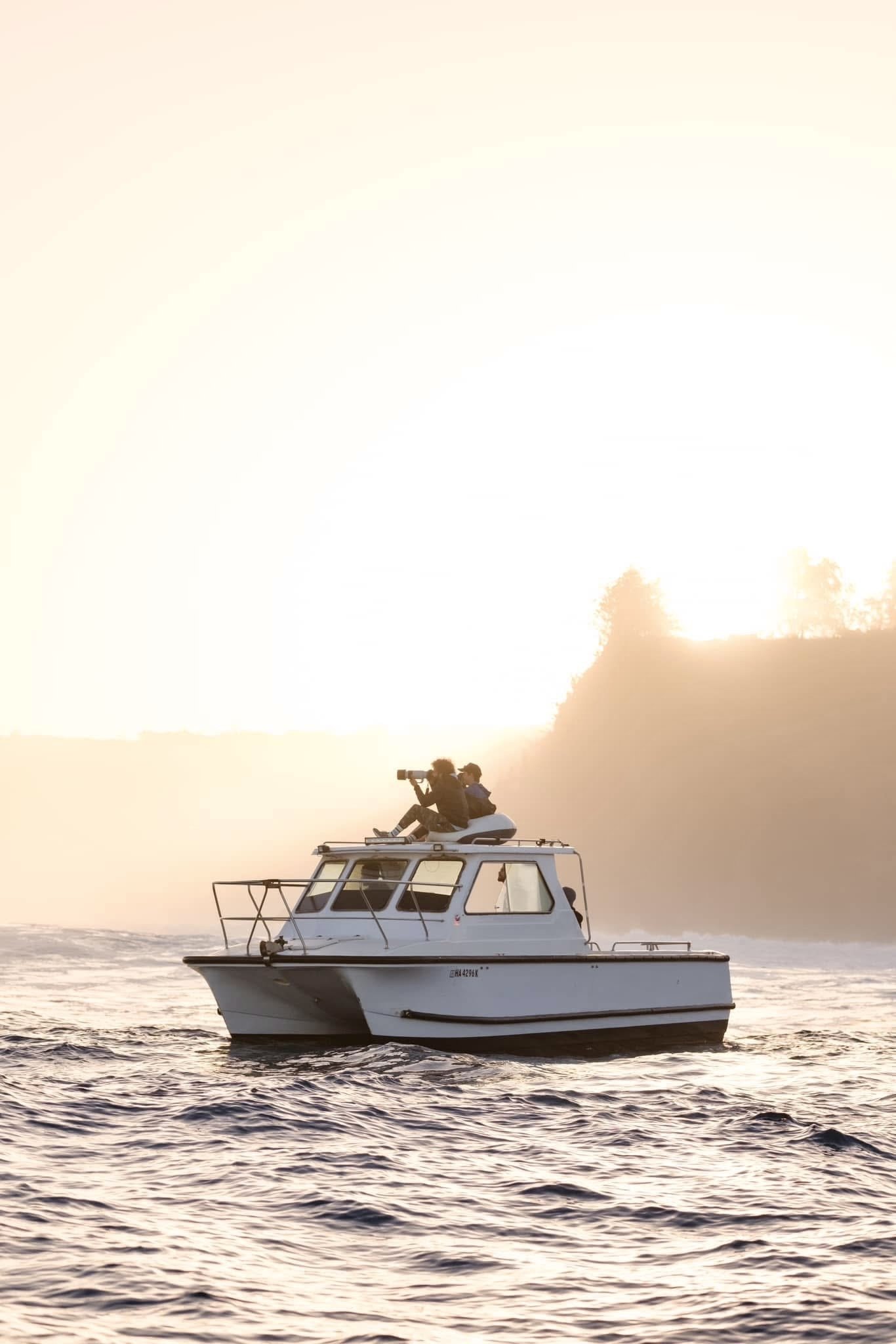 A boat on the water with two people sitting on the roof, one holding a camera, during sunset or sunrise.
