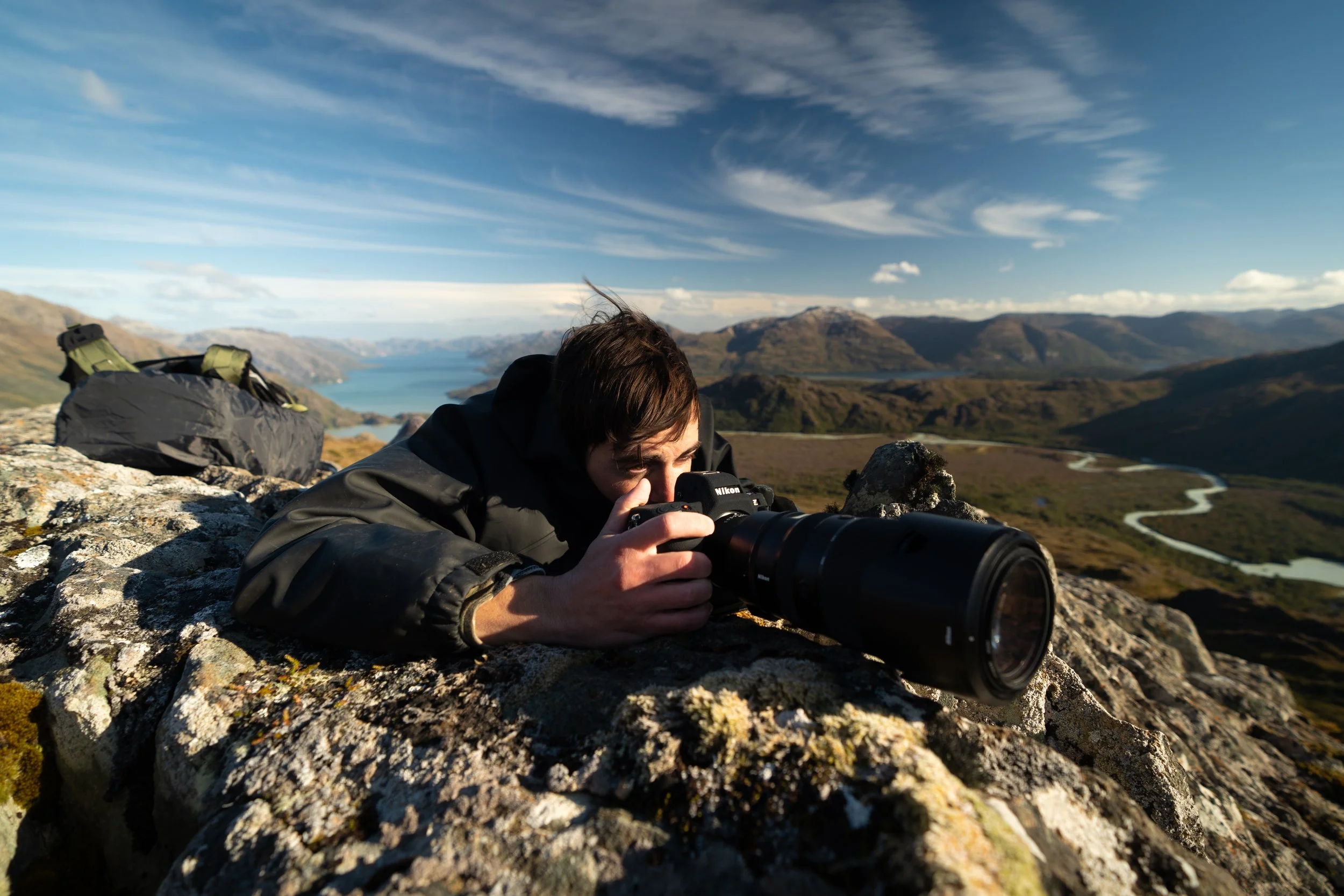 A person lying on a rocky surface outdoors, taking a photograph with a camera, with a scenic landscape of mountains, a lake, and a winding river in the background.