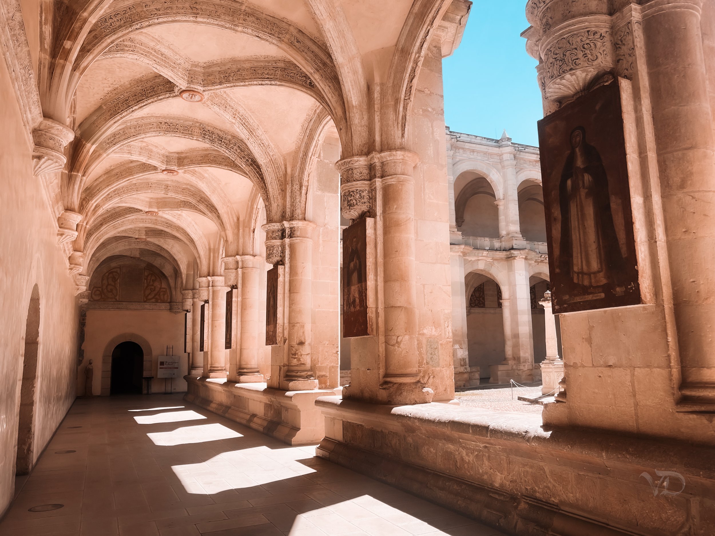 Sunlit corridor in a historic stone building with vaulted ceilings, ornate columns, and religious artwork on display, possibly part of a church or monastery.
