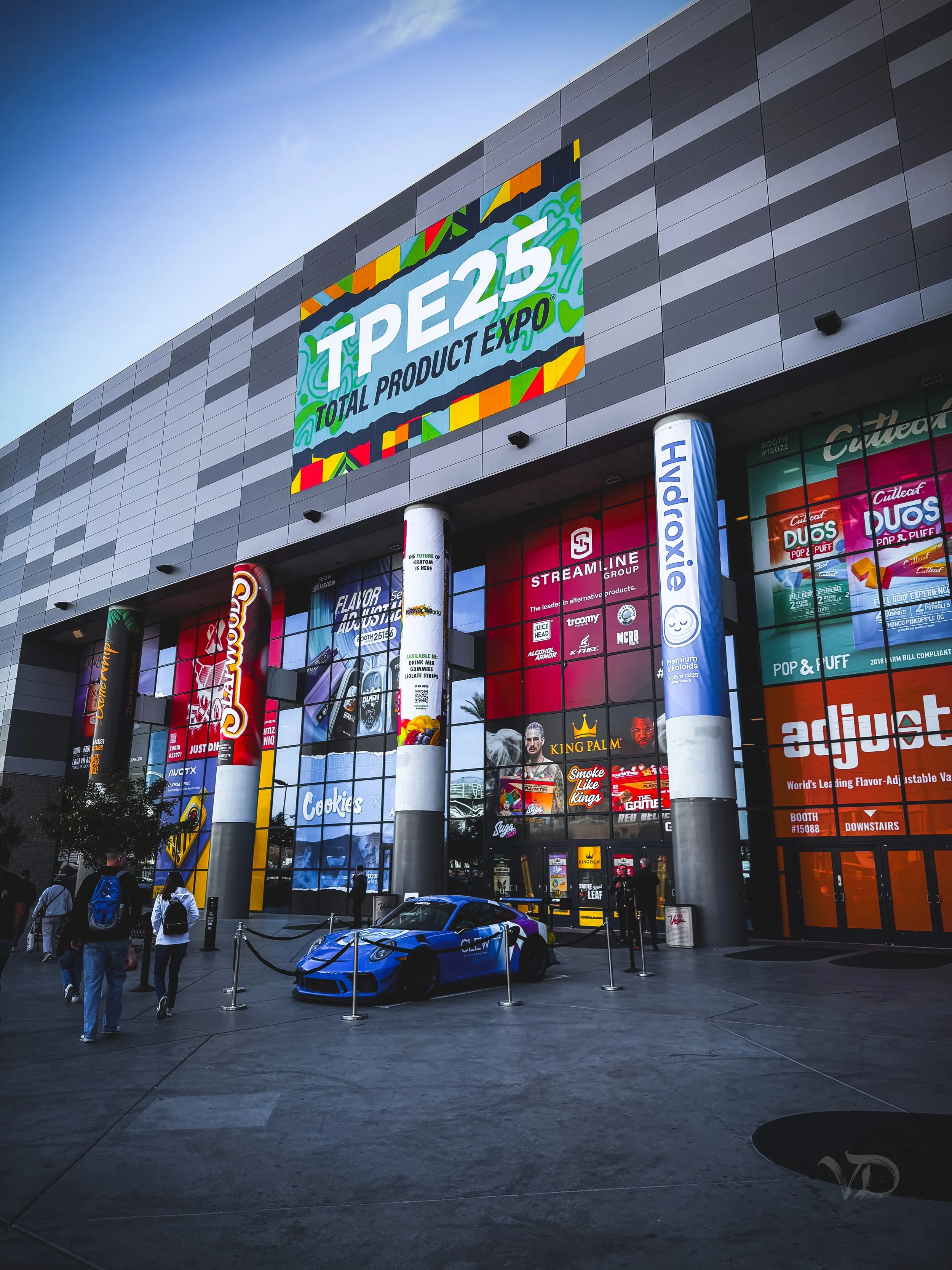 Exterior of the Anaheim Convention Center showcasing banners and advertisements at the Total Product Expo (TPE 25). A blue sports car is displayed in front of the entrance, with several people walking nearby.