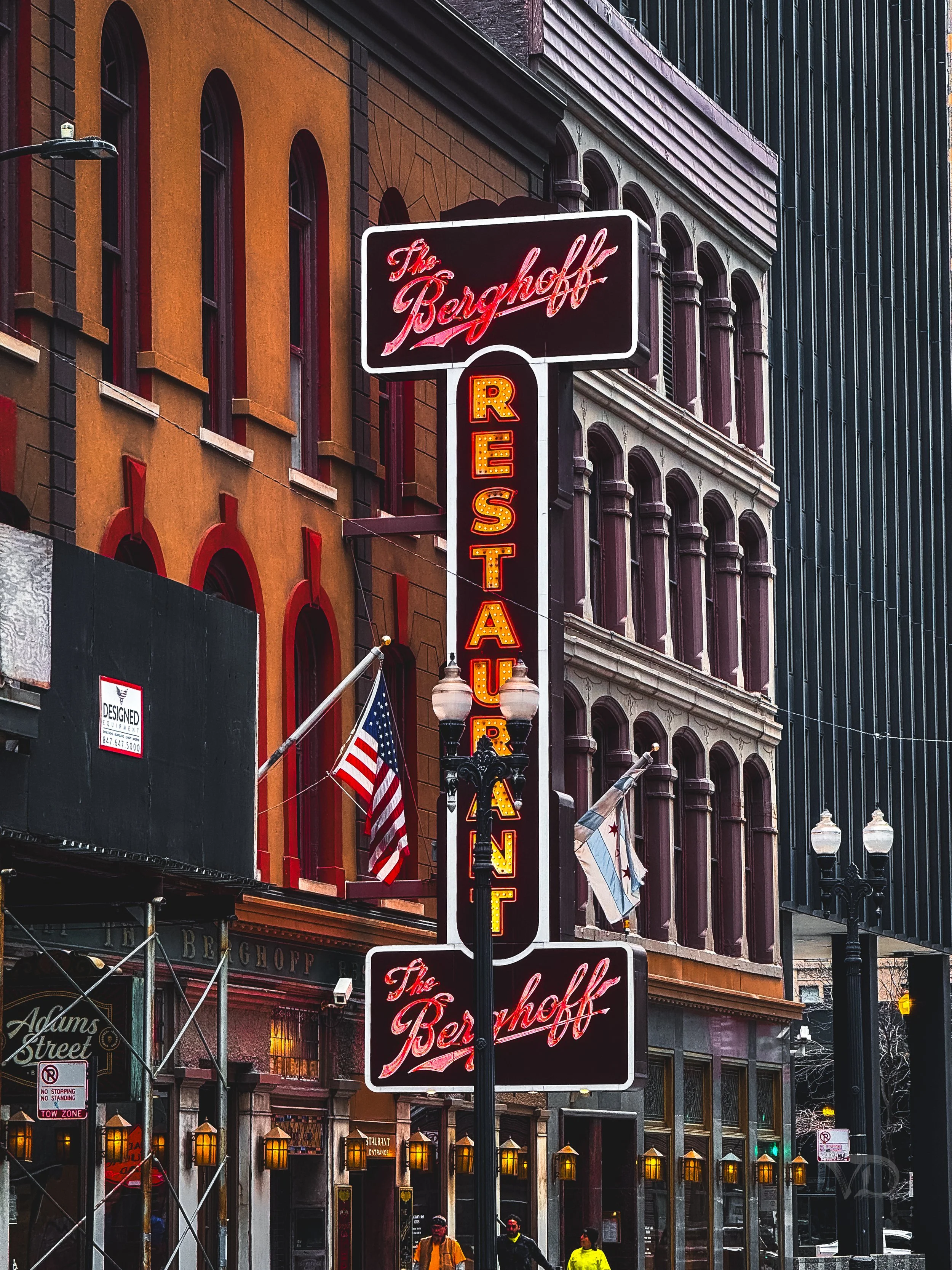 Neon signs for The Bergschff restaurant with a vertical electronic sign that says "RESTAURANT" on a city street, surrounded by buildings with flags, lanterns, and pedestrians.