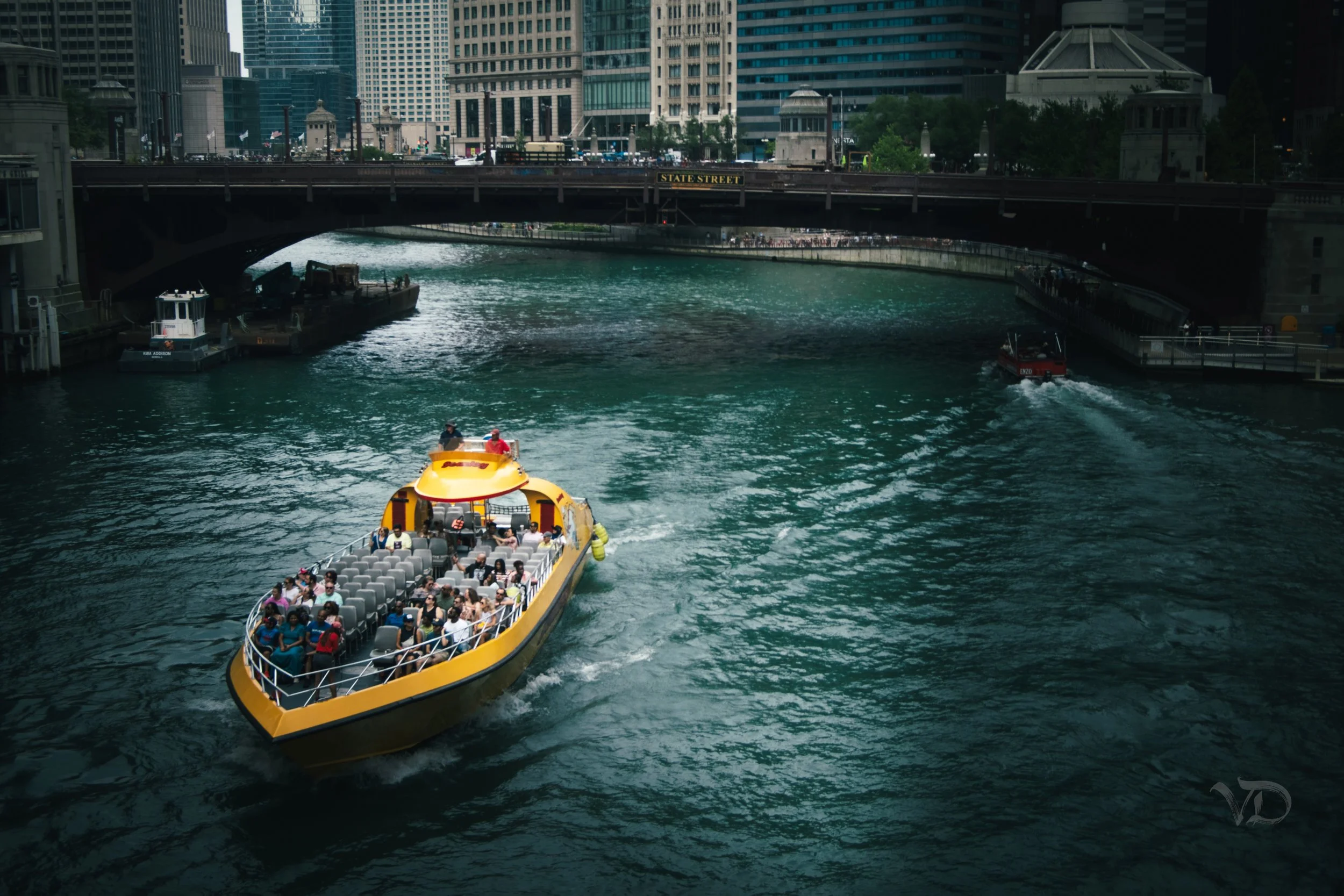 A yellow boat with many passengers on a river in a city, passing under bridges with tall skyscrapers in the background.
