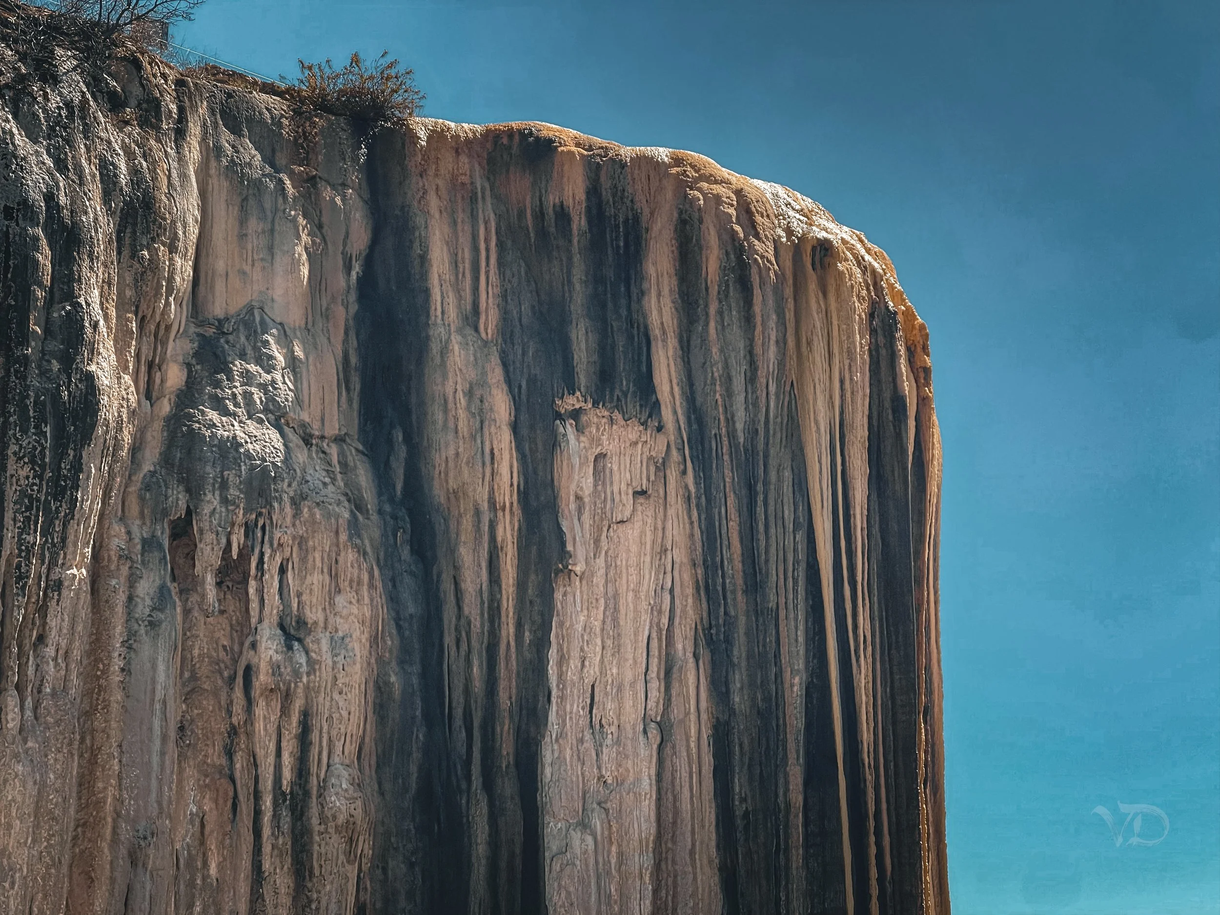 Close-up photograph of a rugged rock formation or cliff with vertical striations, set against a clear blue sky.