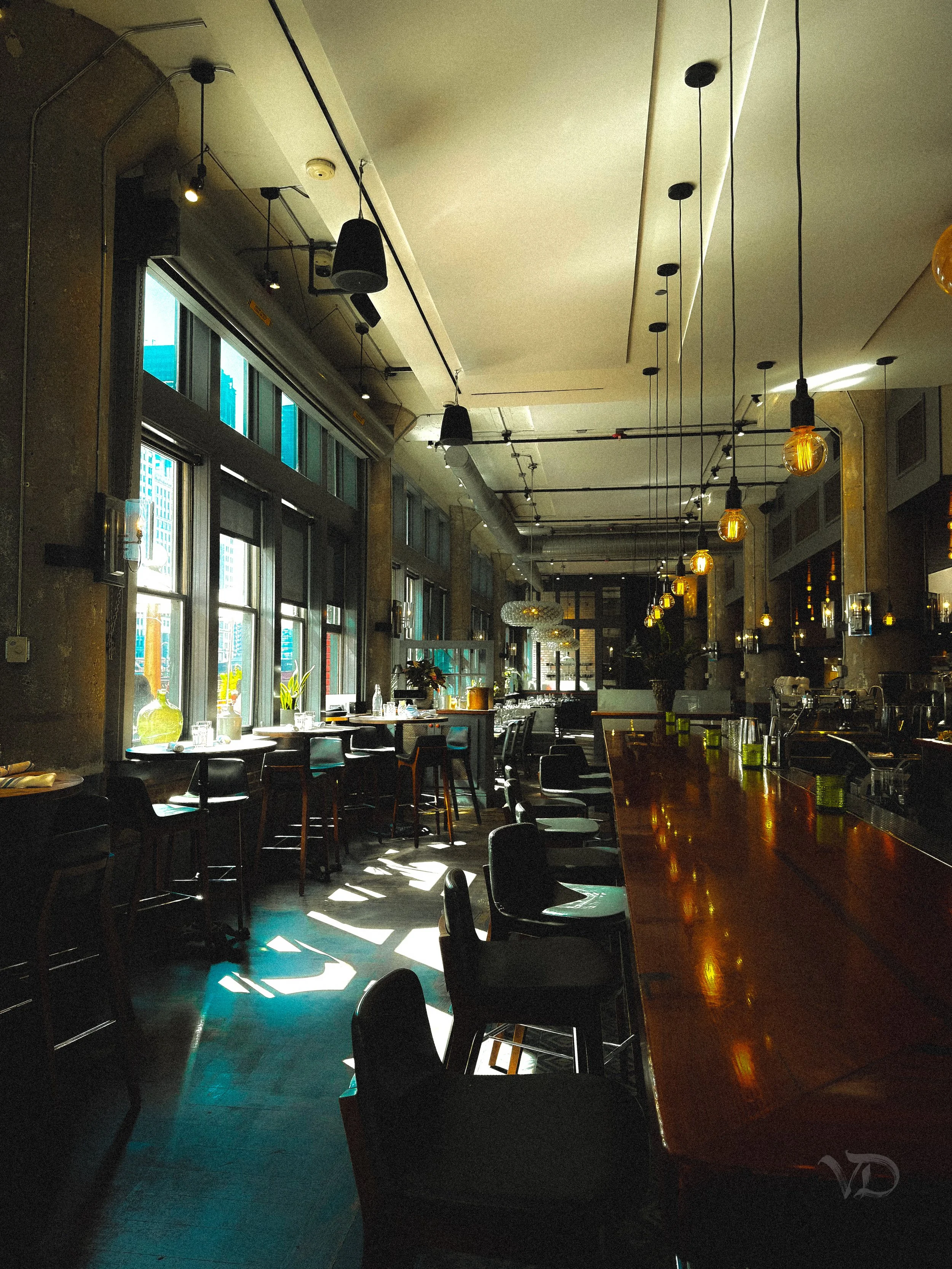 Interior of a modern restaurant or cafe with large windows allowing natural light, dark chairs, a polished wooden bar counter, hanging pendant lights, and some tables with vases and plants.