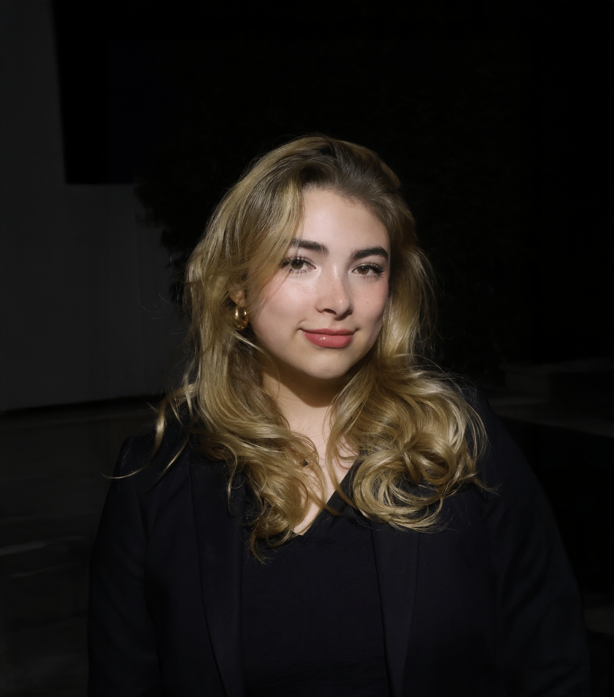 Portrait of a young woman with wavy blonde hair, wearing a black top or blazer, standing in a dimly lit environment with a dark background.