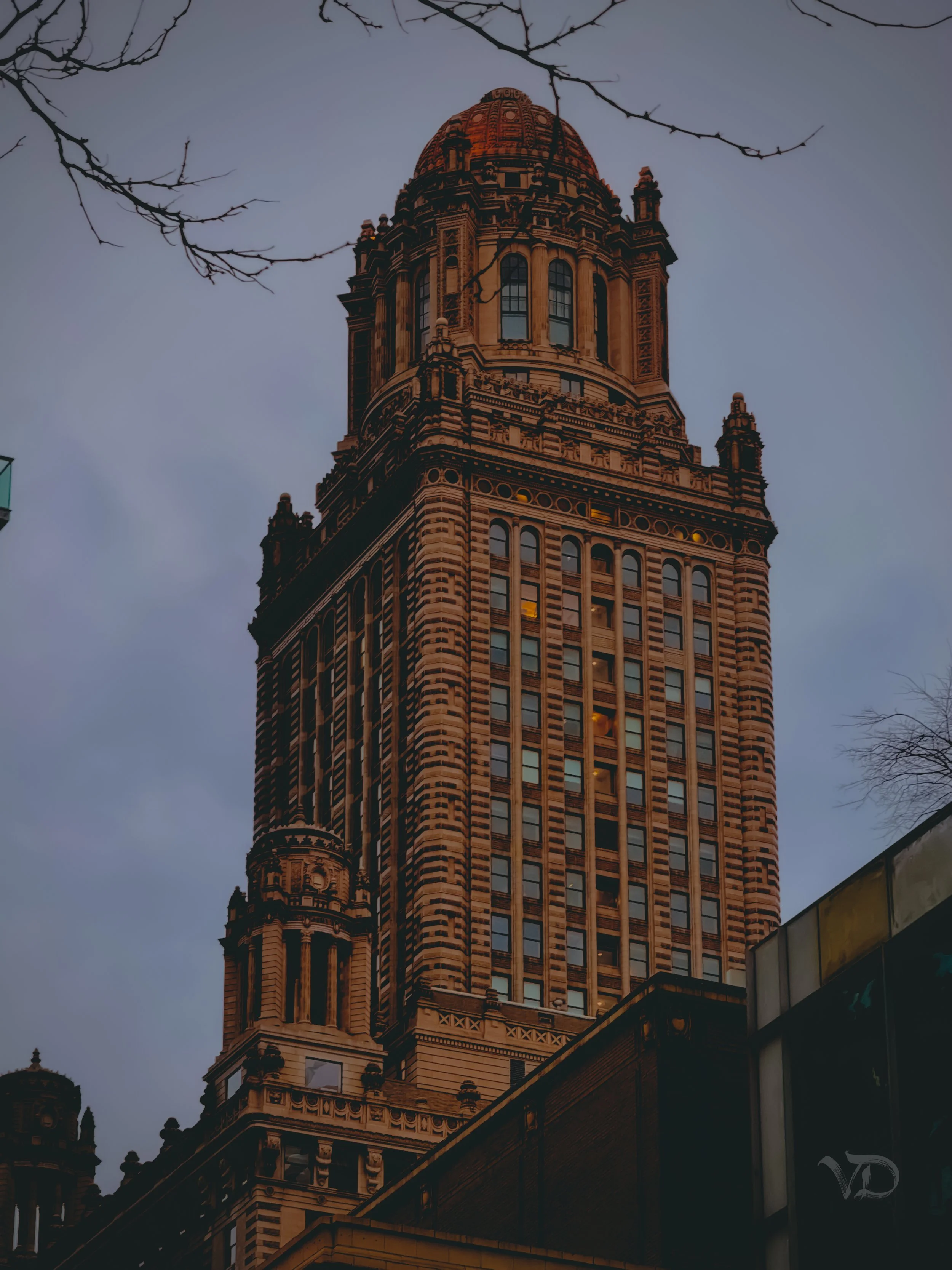 A tall historic skyscraper with intricate architectural details, topped with a dome, against a gray evening sky.