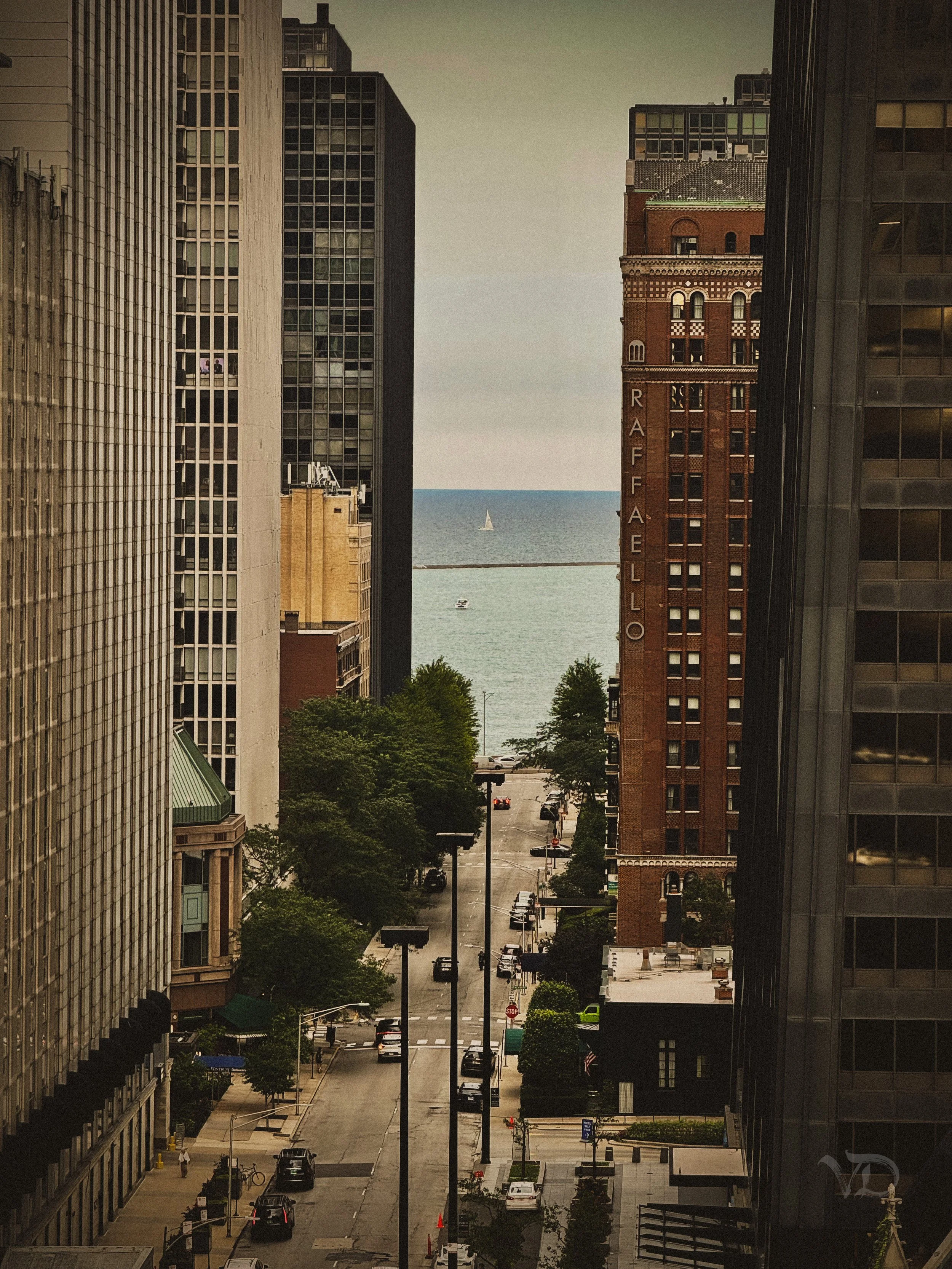 A city street view with tall buildings on each side, leading to a view of a body of water with a sailboat in the distance, and the 