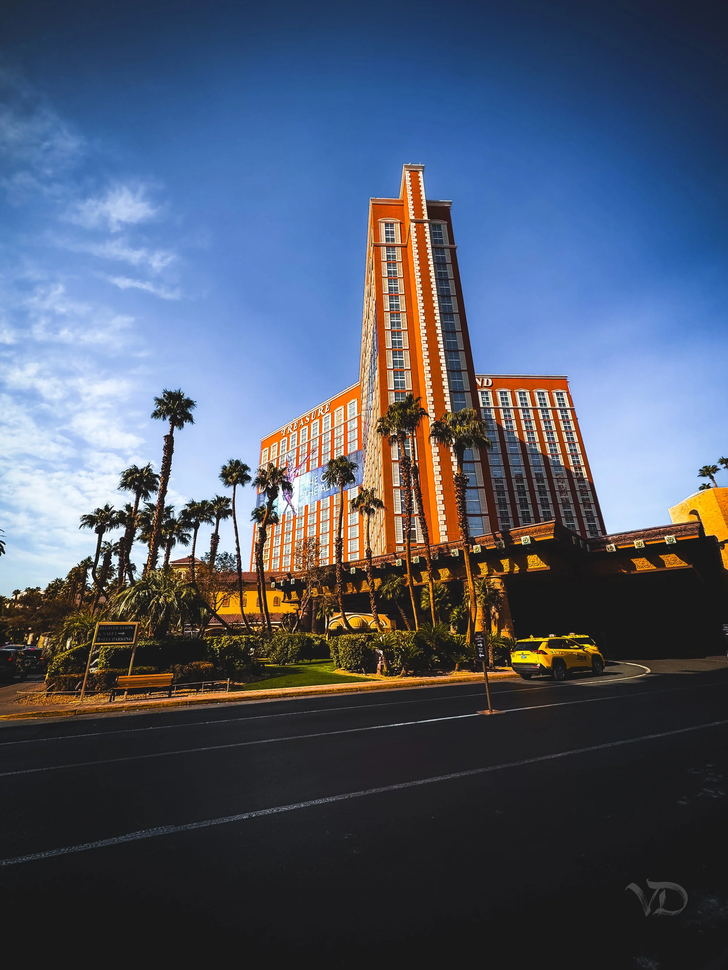 Tall hotel building with red and yellow exterior and palm trees in front, under a blue sky