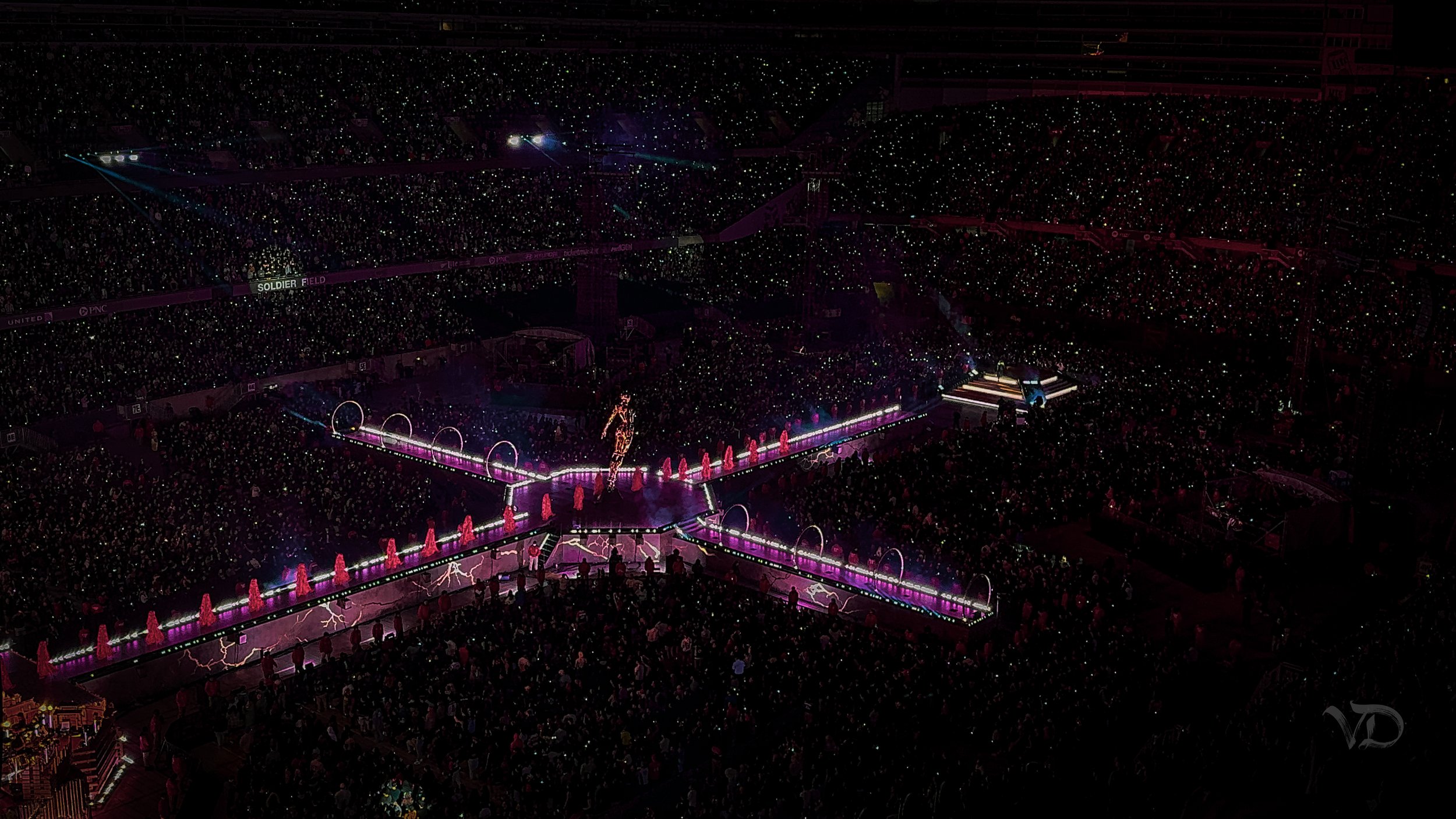 A large indoor stadium filled with a crowd attending a concert or event. The stage features vibrant neon lights and a performer dressed in bright clothing. The arena is illuminated with purple and pink lighting, creating a lively atmosphere.