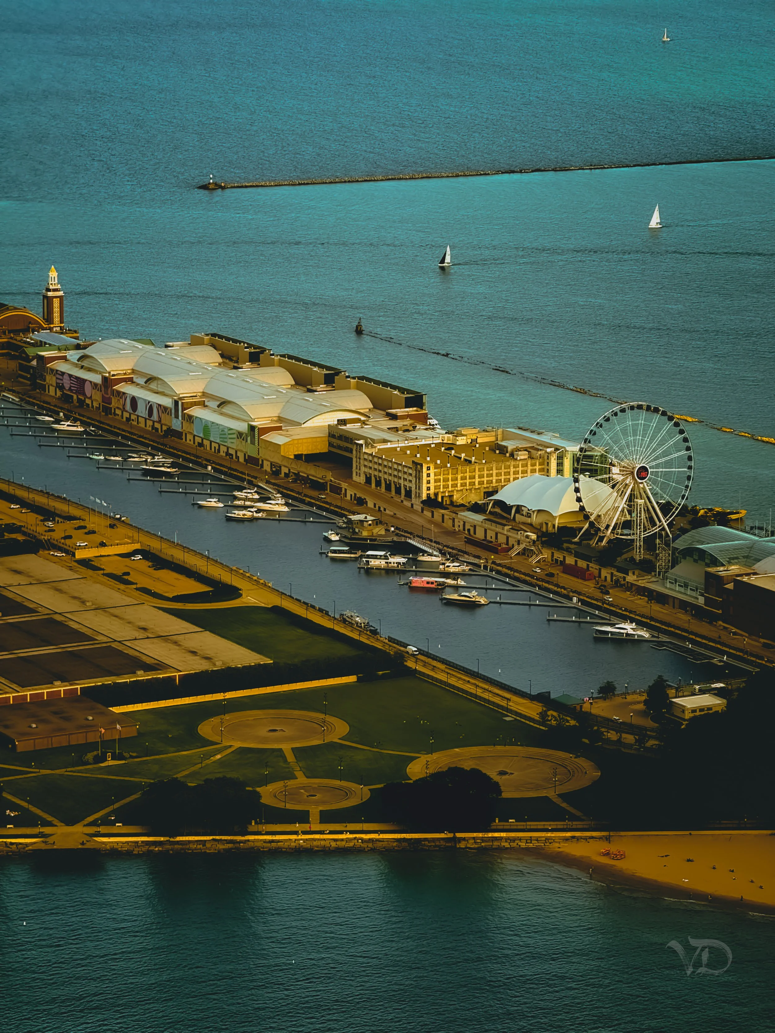 An aerial view of a waterfront with a carnival-style Ferris wheel, boats docked along a pier, a large building with a colorful exterior, and sailboats on the water. Green park area with circular pathways is visible in the foreground.