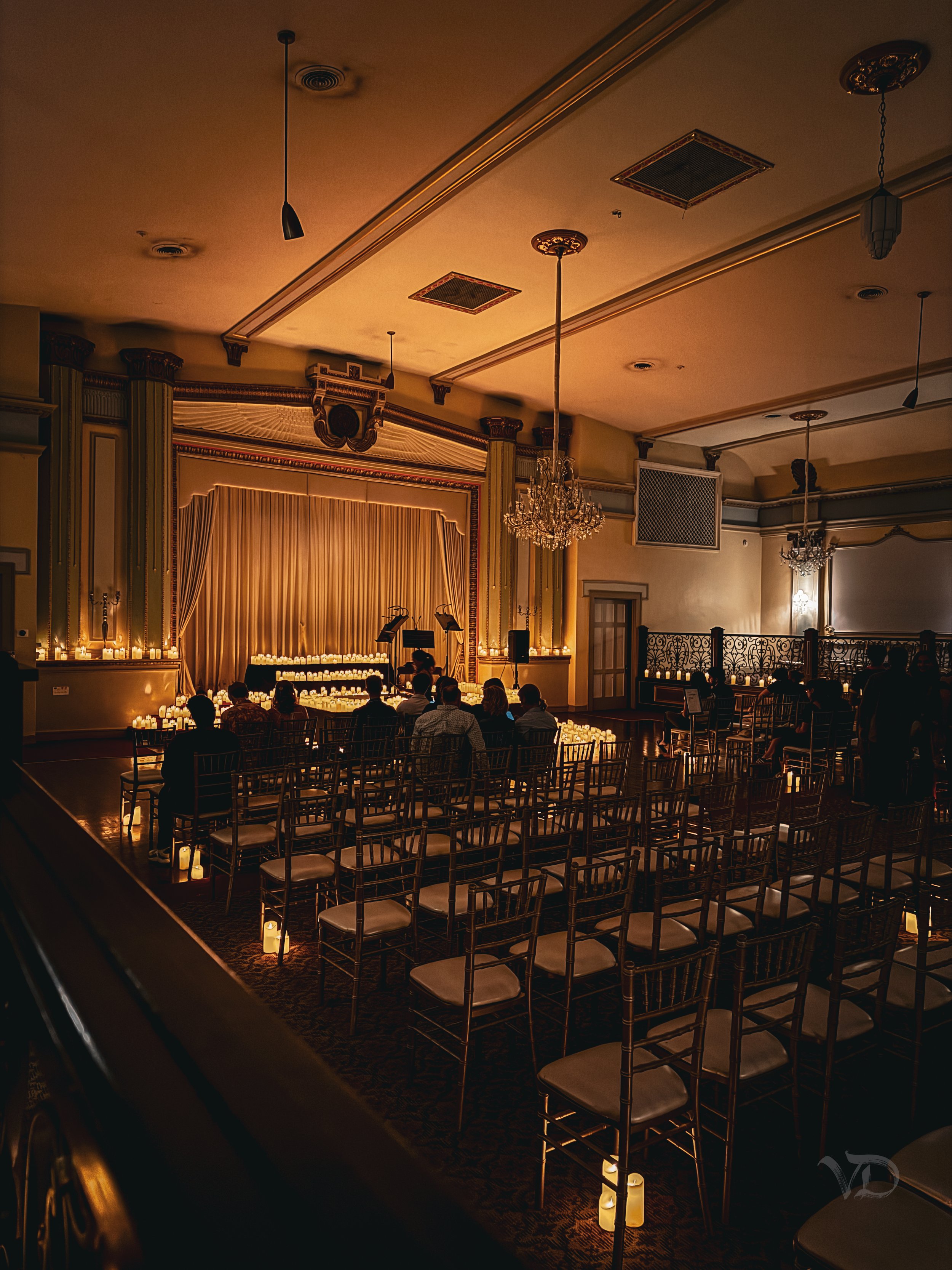 Elegant dimly lit event venue with a stage, chandelier, and rows of chairs surrounded by candles, set up for a performance or ceremony