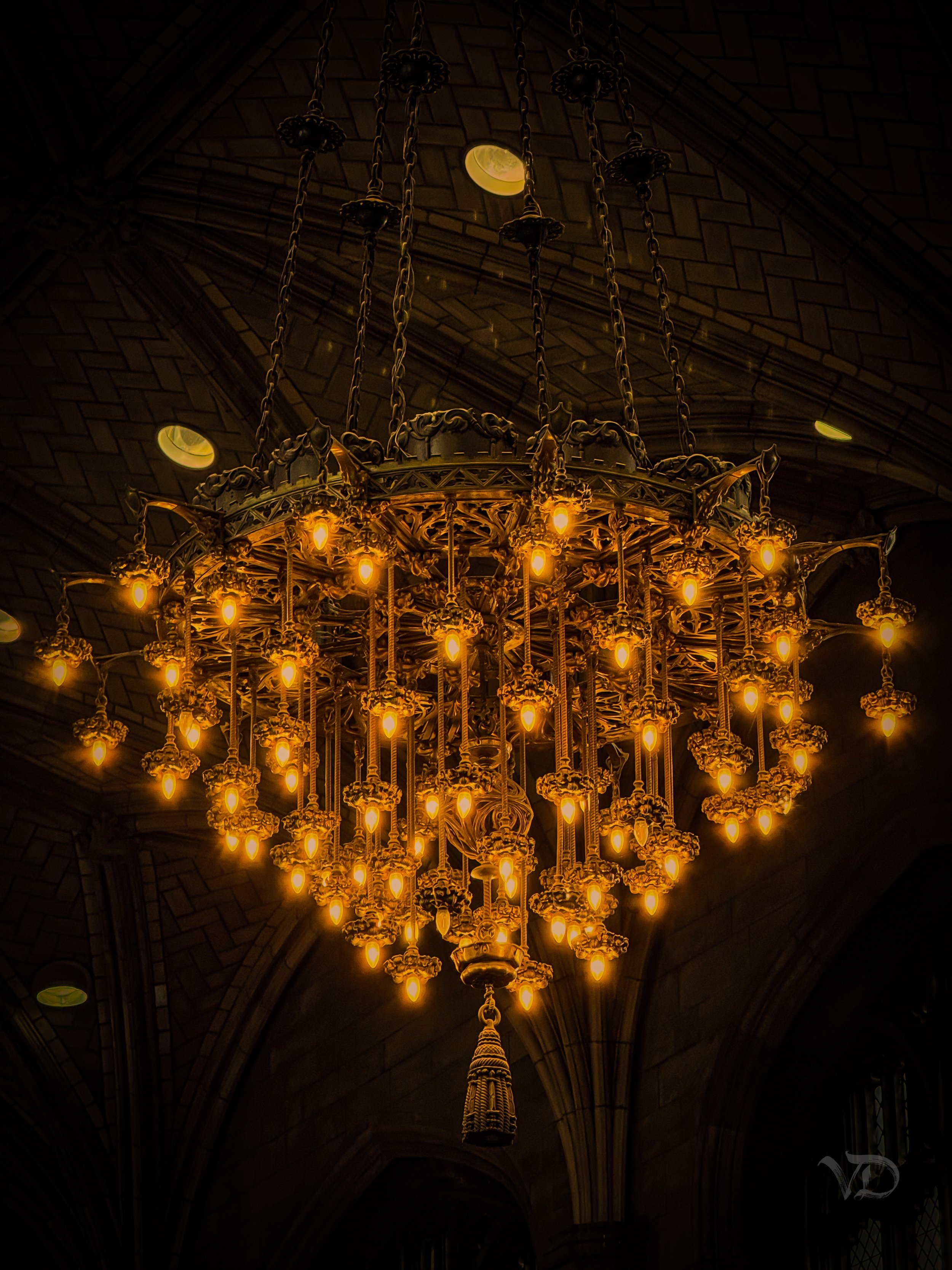 A large ornate chandelier with multiple lit bulbs hanging from a vaulted stone ceiling.