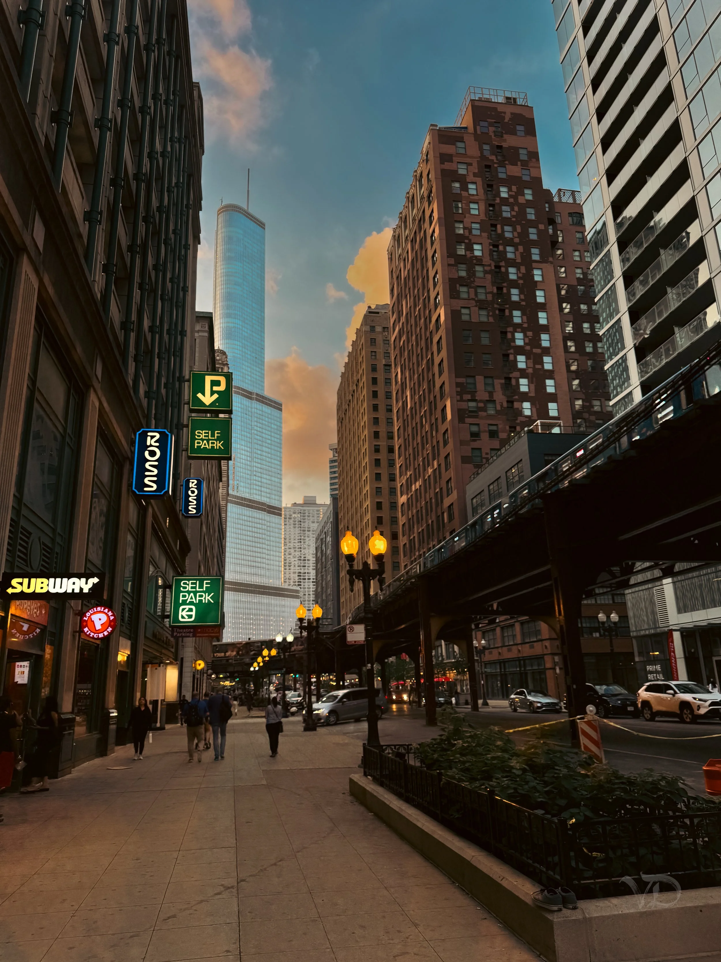 City street scene at sunset with tall skyscrapers, retail stores including Subway, parked cars, pedestrians walking, street lamps, and an elevated train track in downtown Chicago.