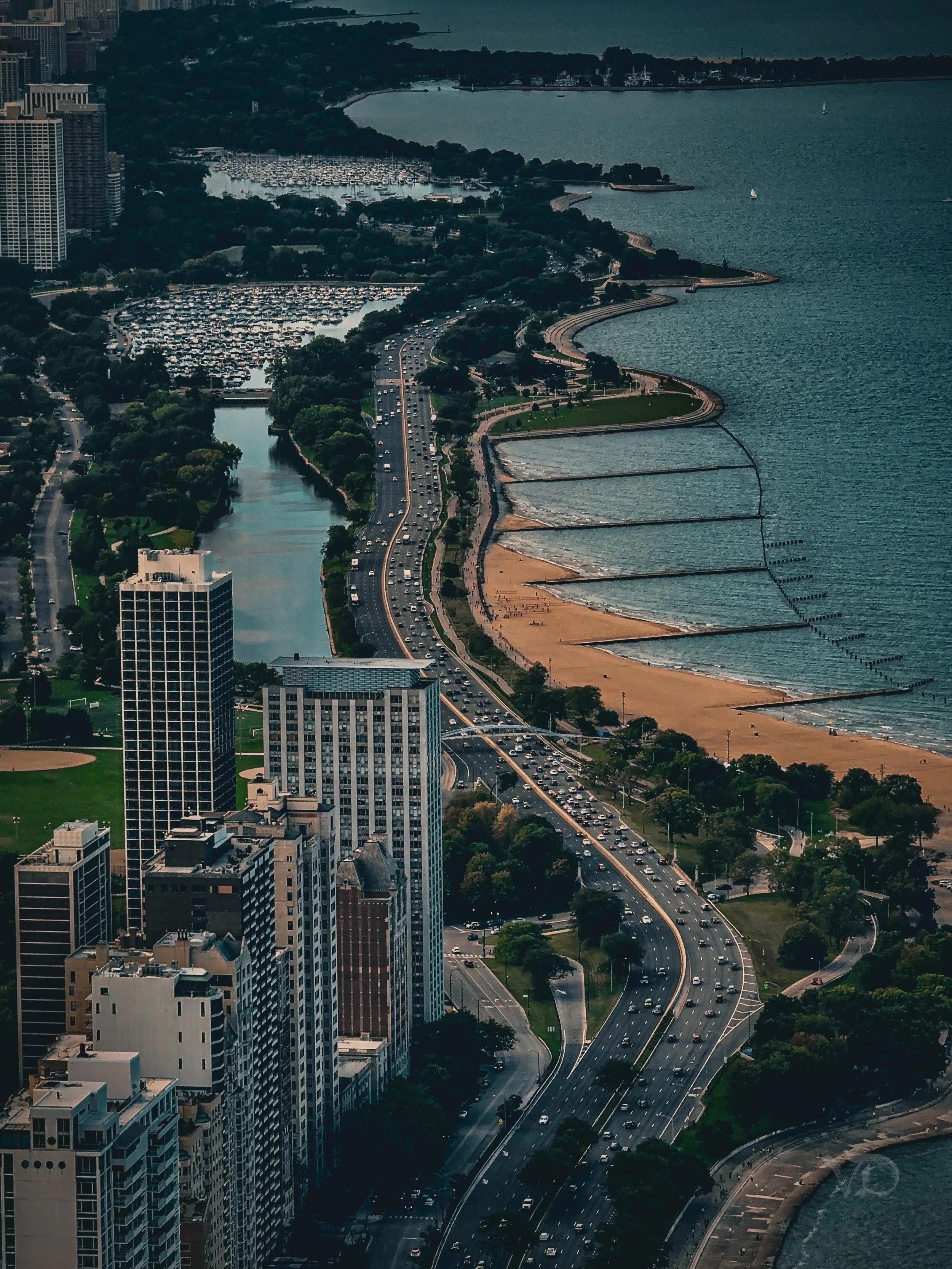 Aerial view of a cityscape with a highway running along a lake, high-rise buildings, parks, and a marina with boats on the water.