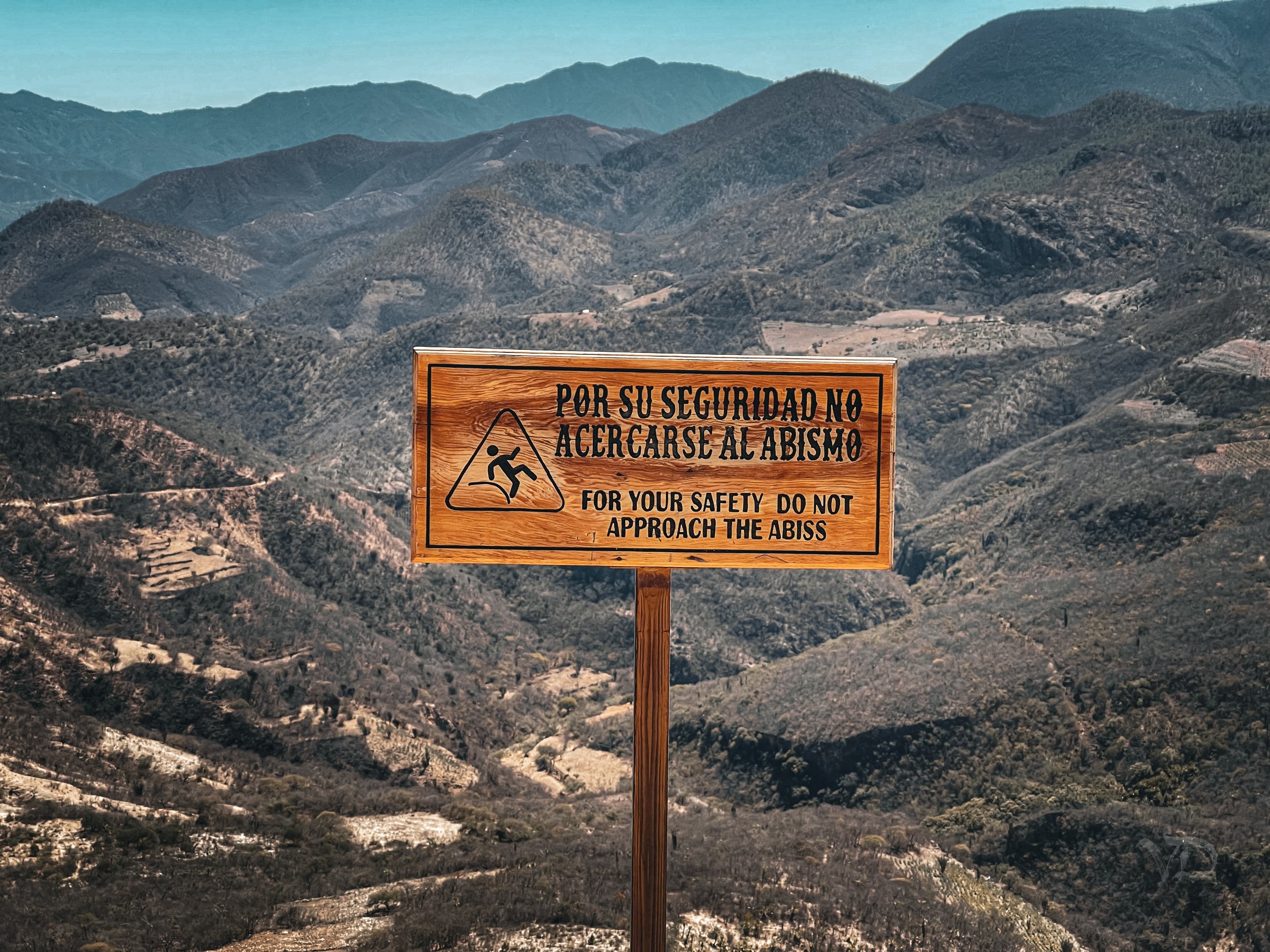 Wooden warning sign in Spanish and English reading, 'For your safety do not approach the abyss,' with a pictogram of a person falling, set against a mountainous landscape.