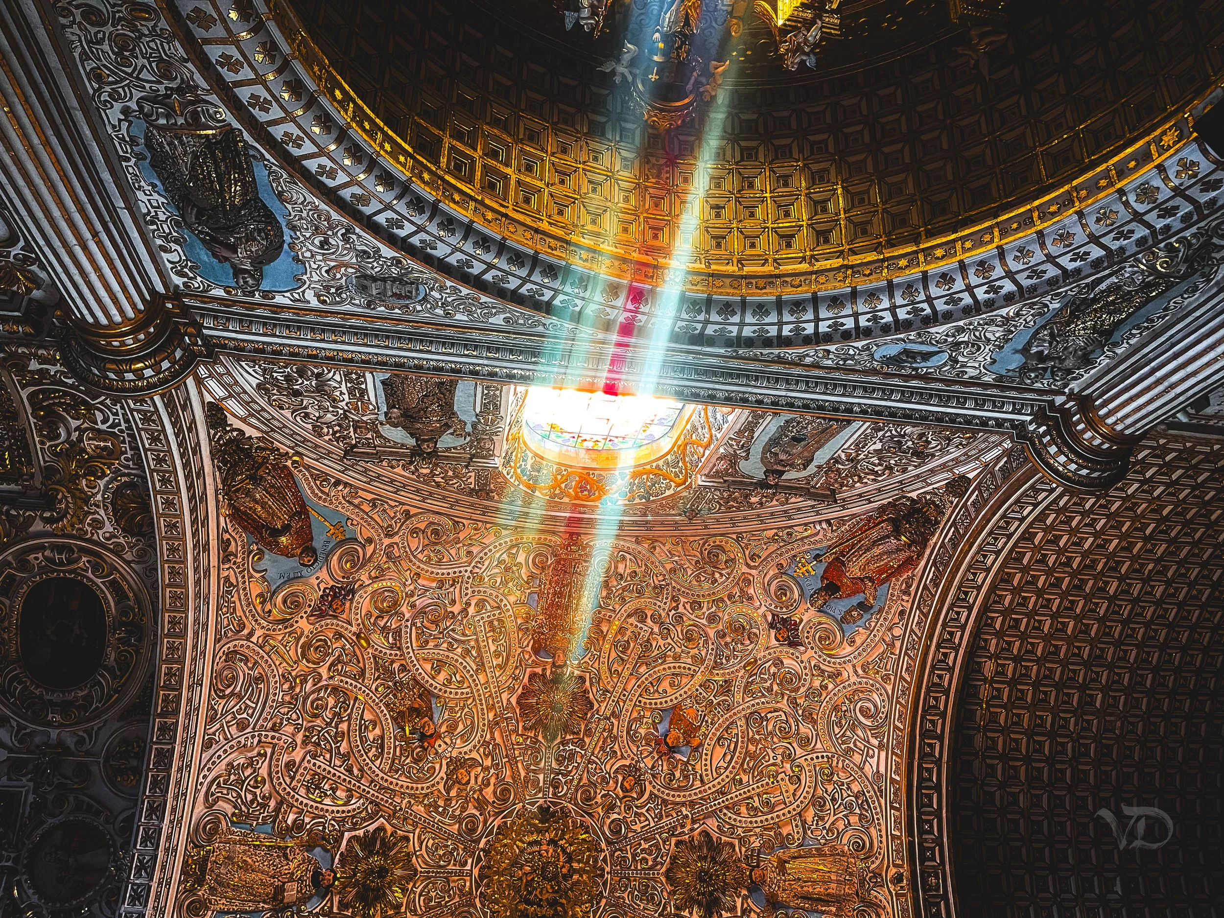 Sunlight streaming through a stained glass skylight onto an ornate, gold and silver decorated ceiling with intricate patterns and sculptures.