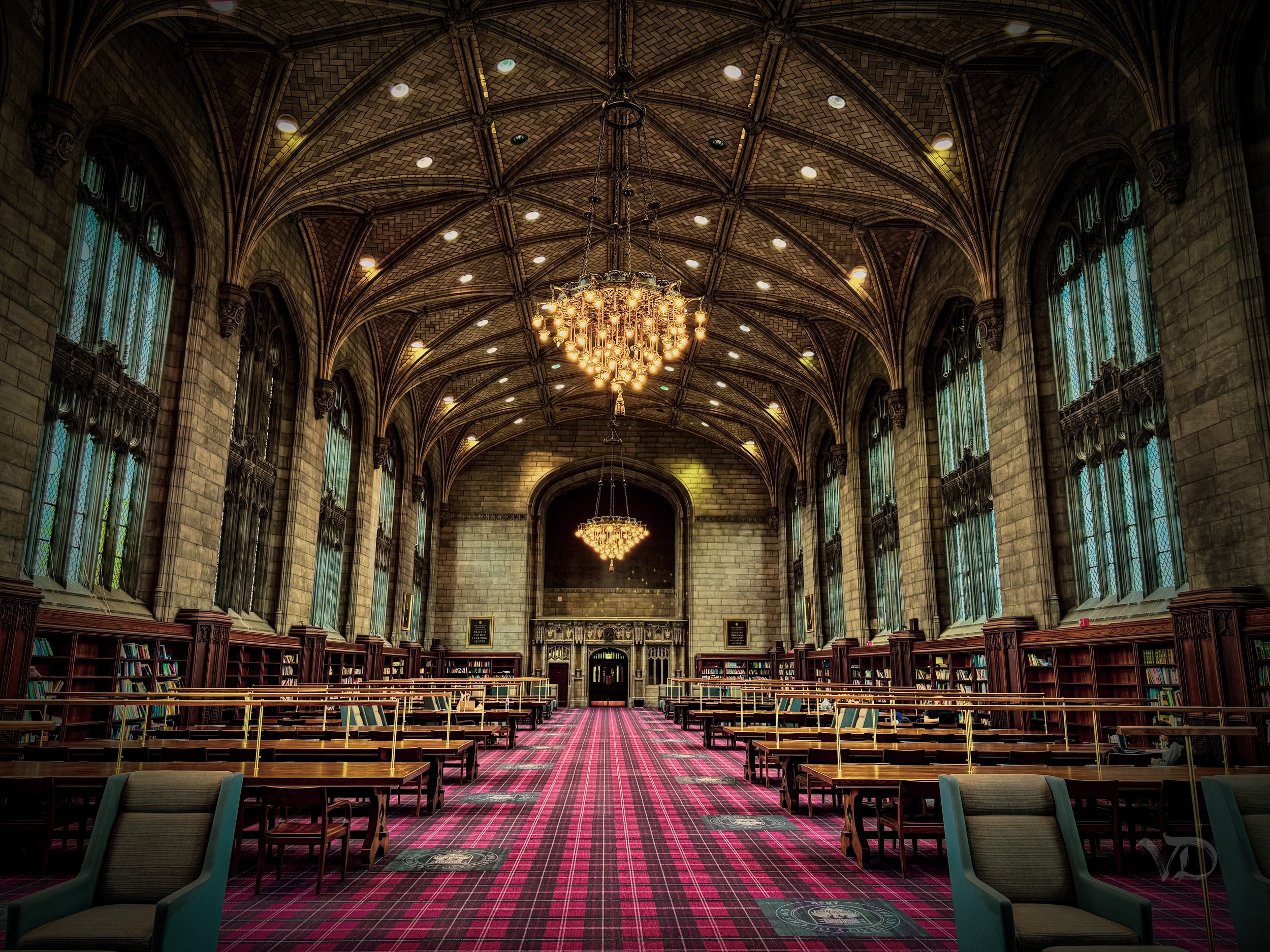 Interior of a historic library with high vaulted ceilings, large stained glass windows, chandeliers hanging from the ceiling, and rows of bookshelves along the walls. There are tables and chairs arranged for reading or studying. The carpet is pattern