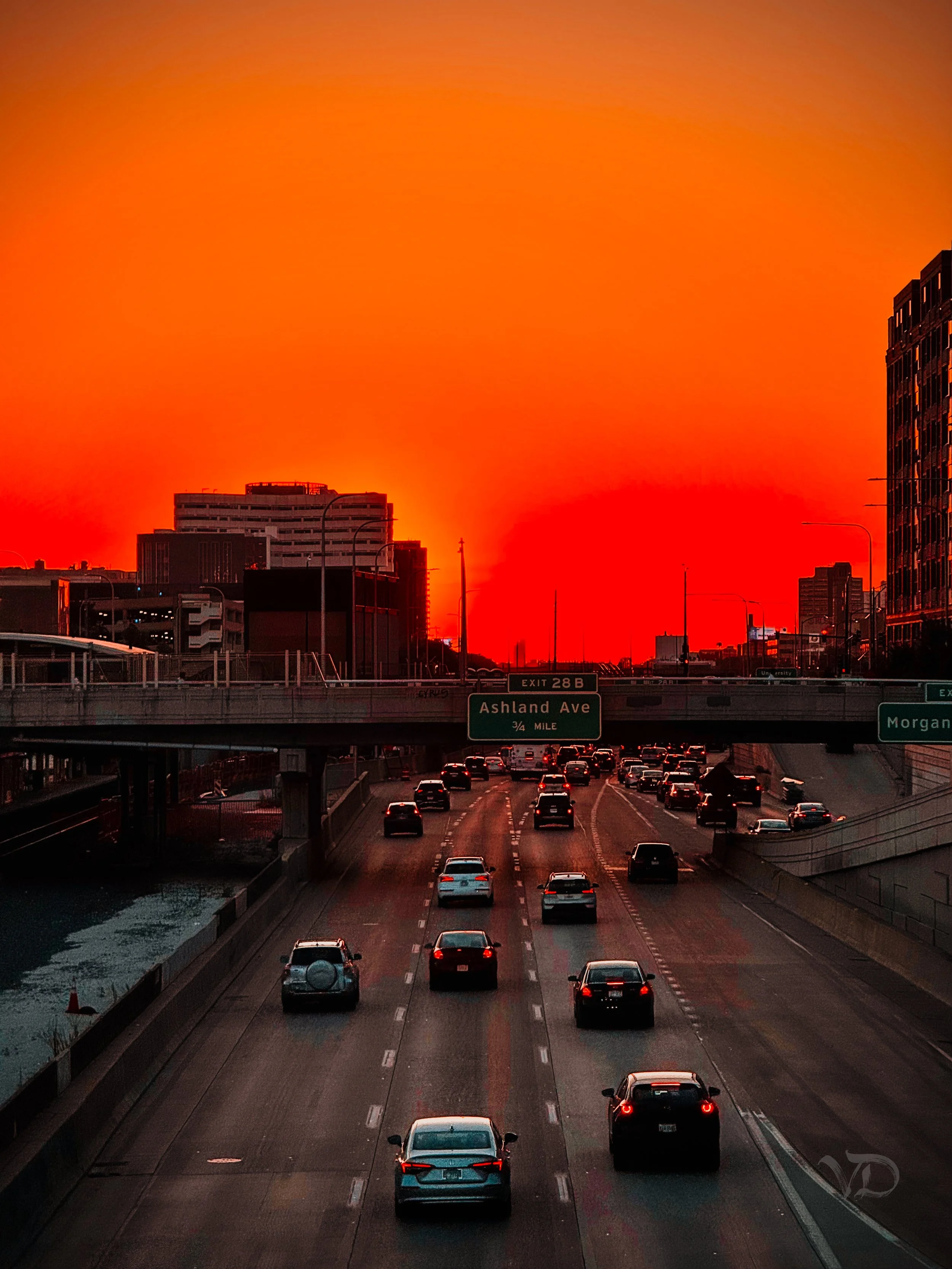 City street scene during a vivid sunset with orange sky, high traffic, and tall buildings in the background.
